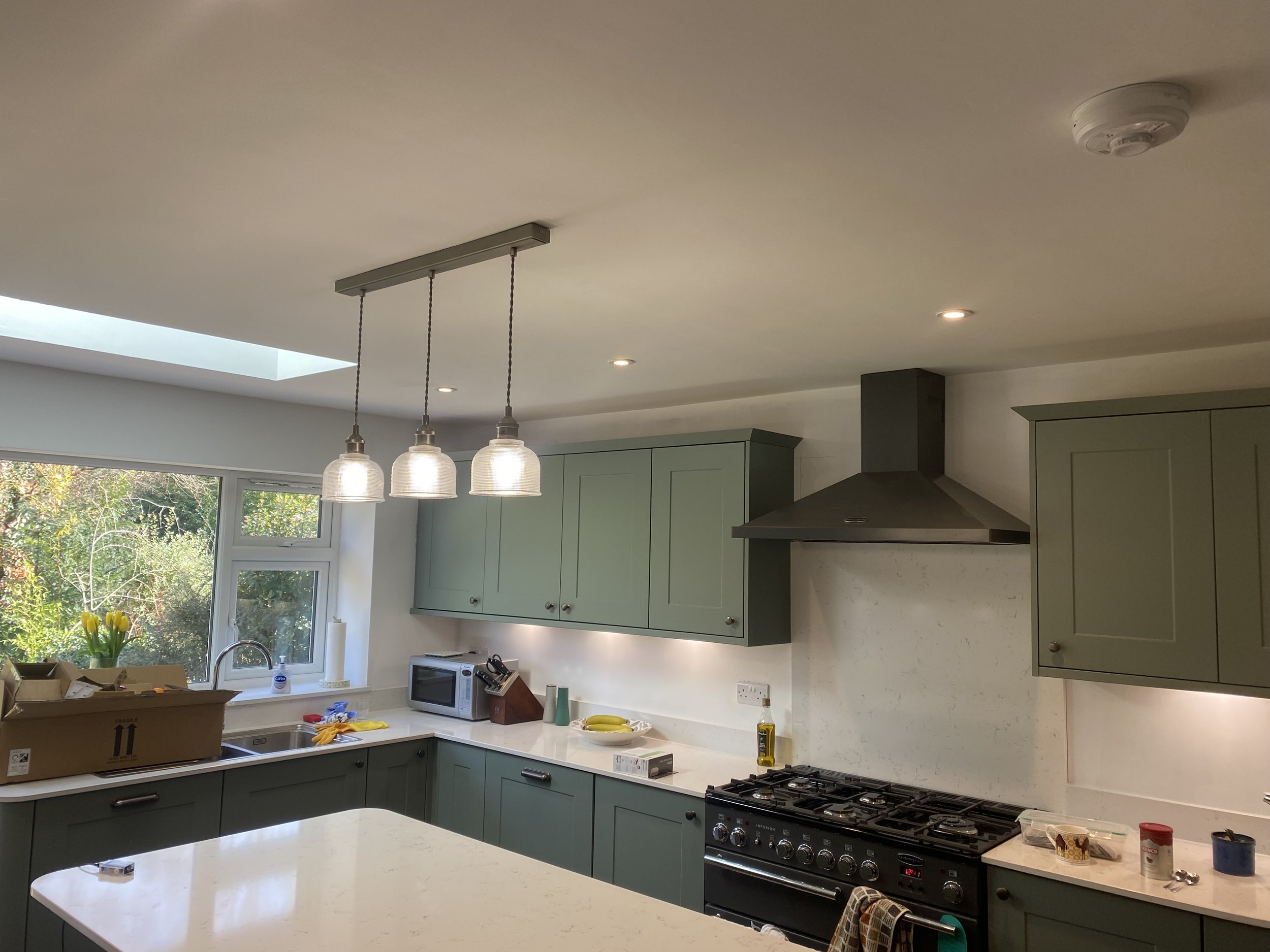 A modern kitchen with light green cabinets, a white countertop, and a gas stove. Several items are on the counter, including a microwave, bananas, and a box. There are large windows and a skylight providing natural light.