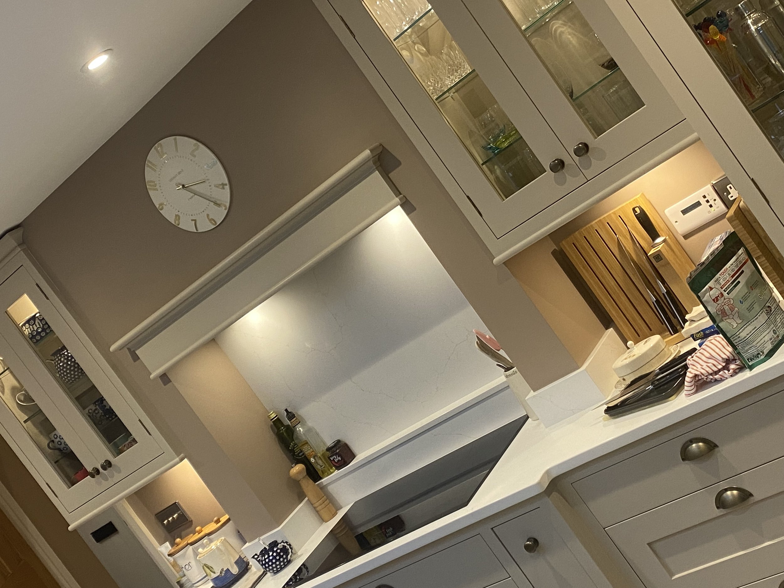 A kitchen with white cabinets and a glass-front owl cabinet. A white clock on the wall, various bottles and jars on the counter, a cutting board, and a mixing bowl with a striped cloth are visible on the kitchen island.