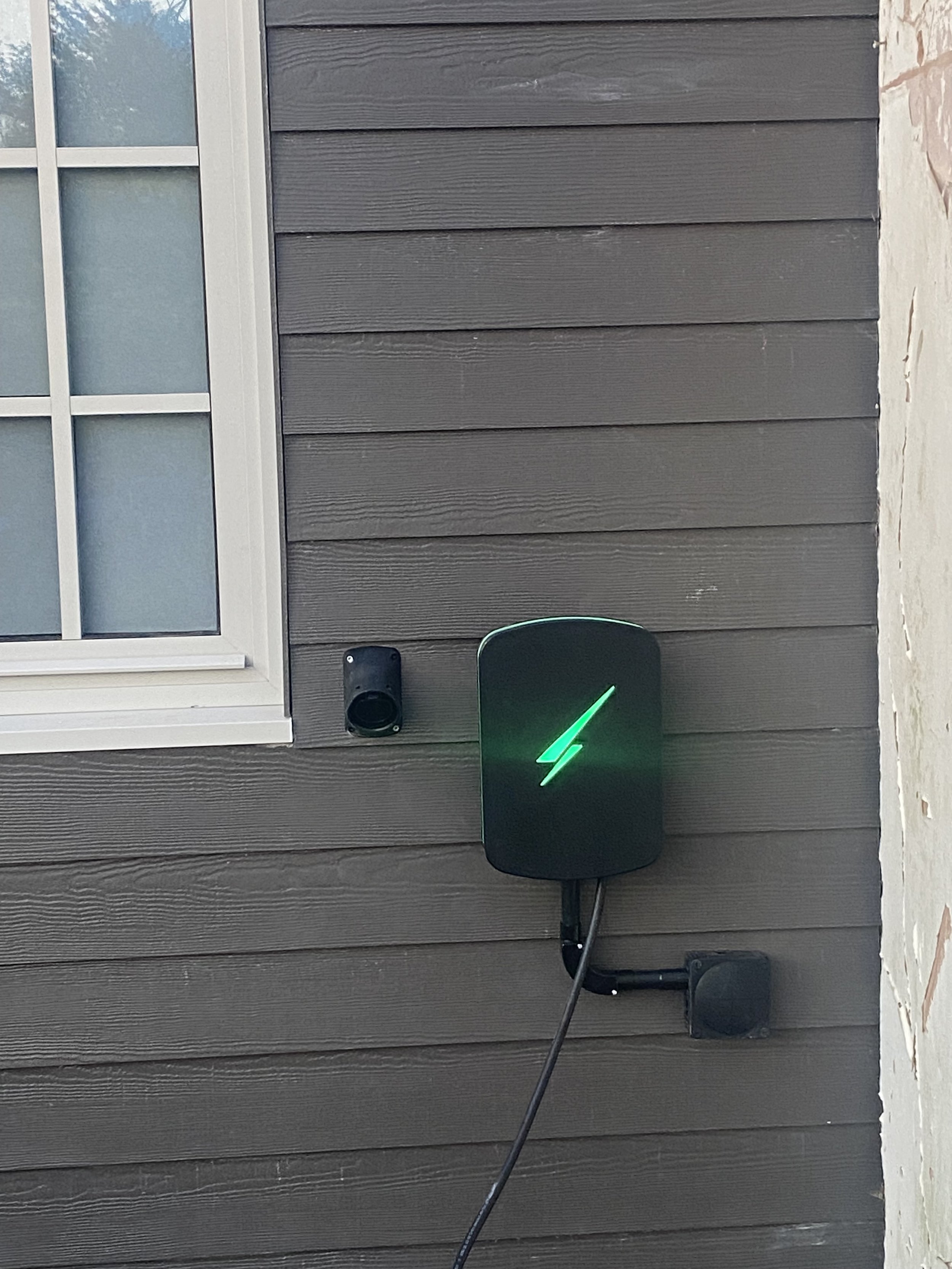 A black electric vehicle charging station with a green lightning bolt symbol, mounted on a dark gray wooden wall, next to a black speaker or sensor and a power outlet, with part of a window on the left side.