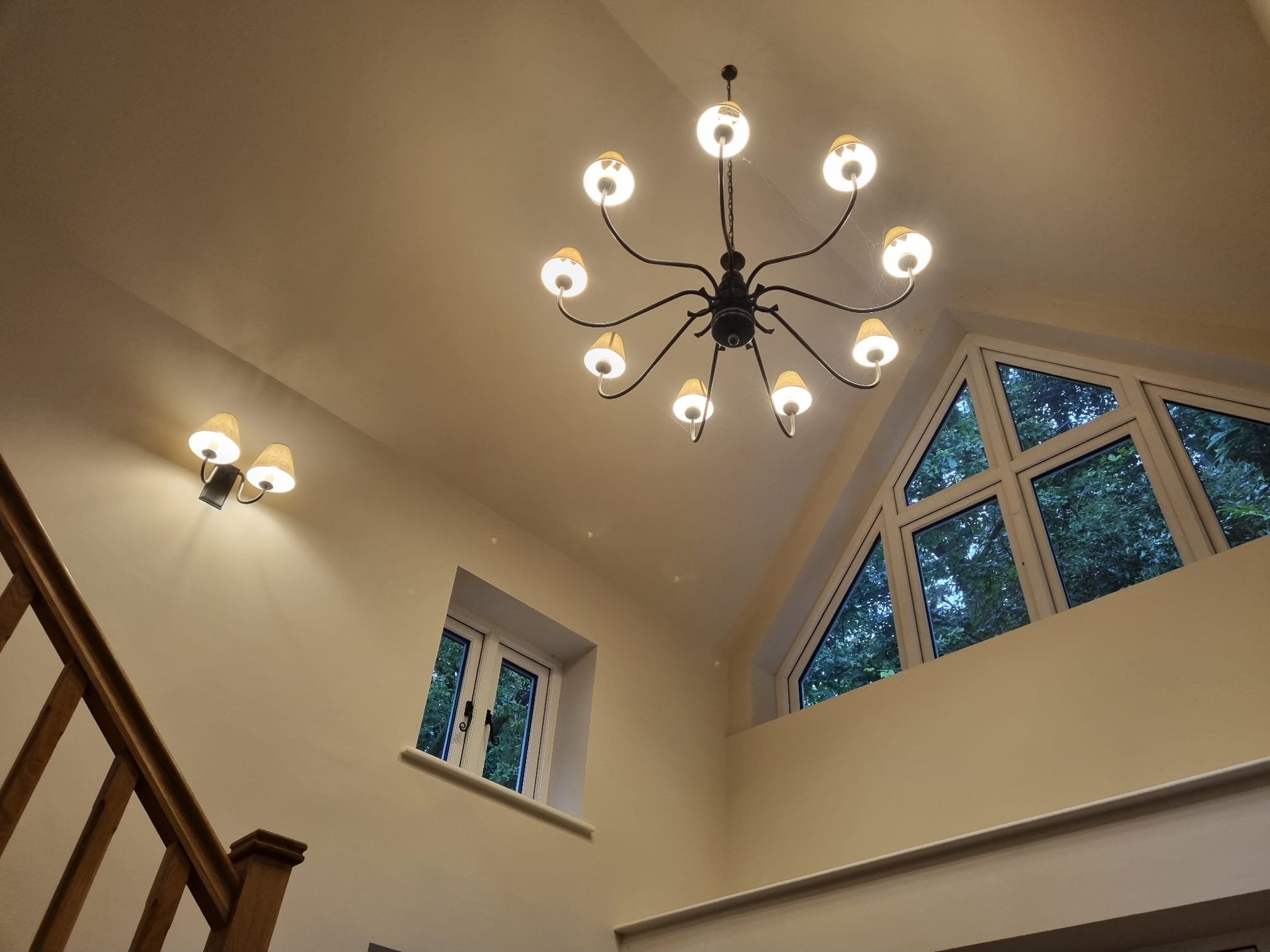 A chandelier with multiple arms and lampshades hanging from a ceiling. A wall with a light fixture, a staircase, and large windows showing trees outside are visible.