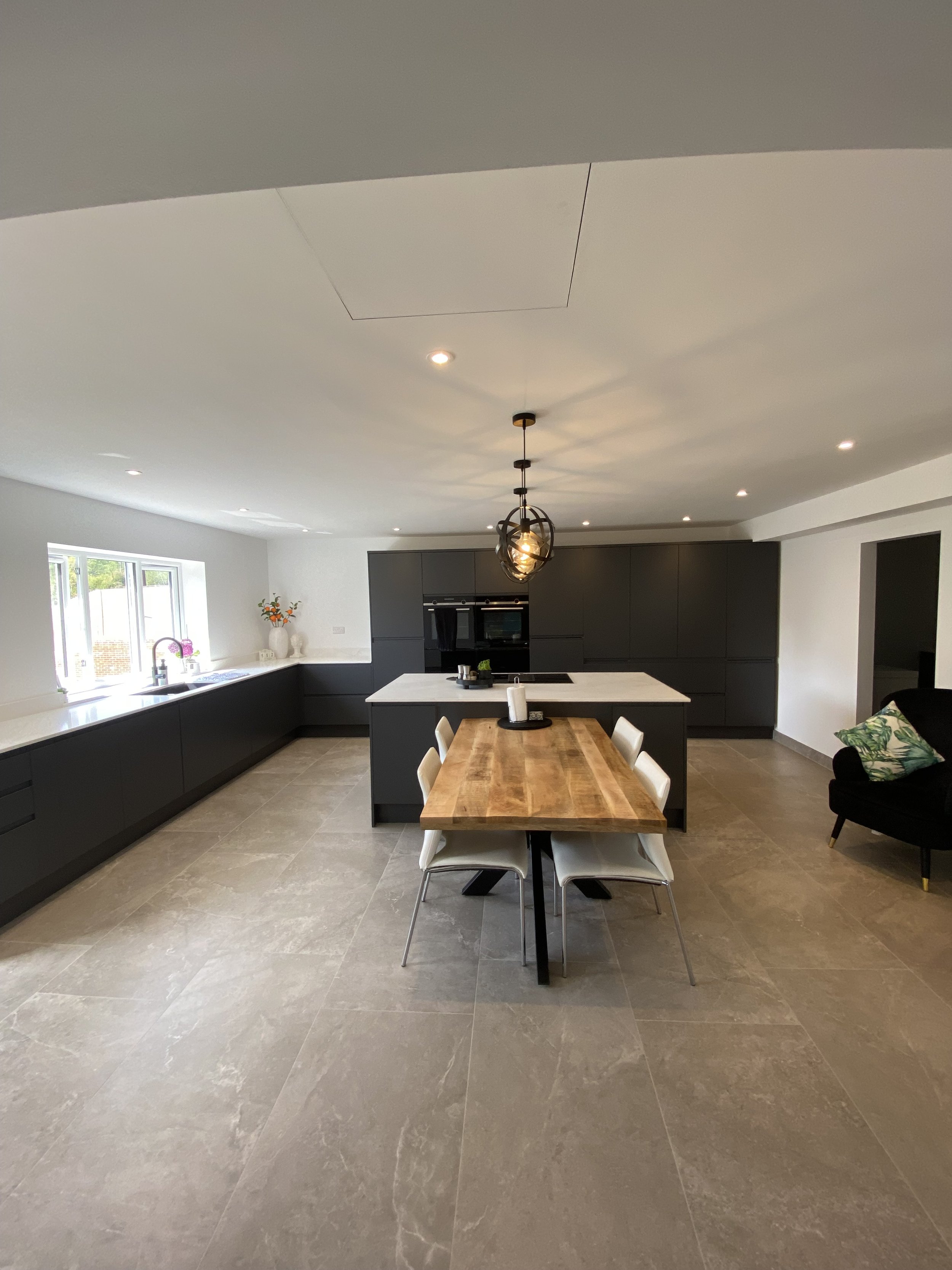 A modern open-concept kitchen and dining area with a large window, black cabinets, a white countertop, a wooden dining table with six chairs, and a light fixture hanging above the table. There is a black sofa with a green pillow on the right side and