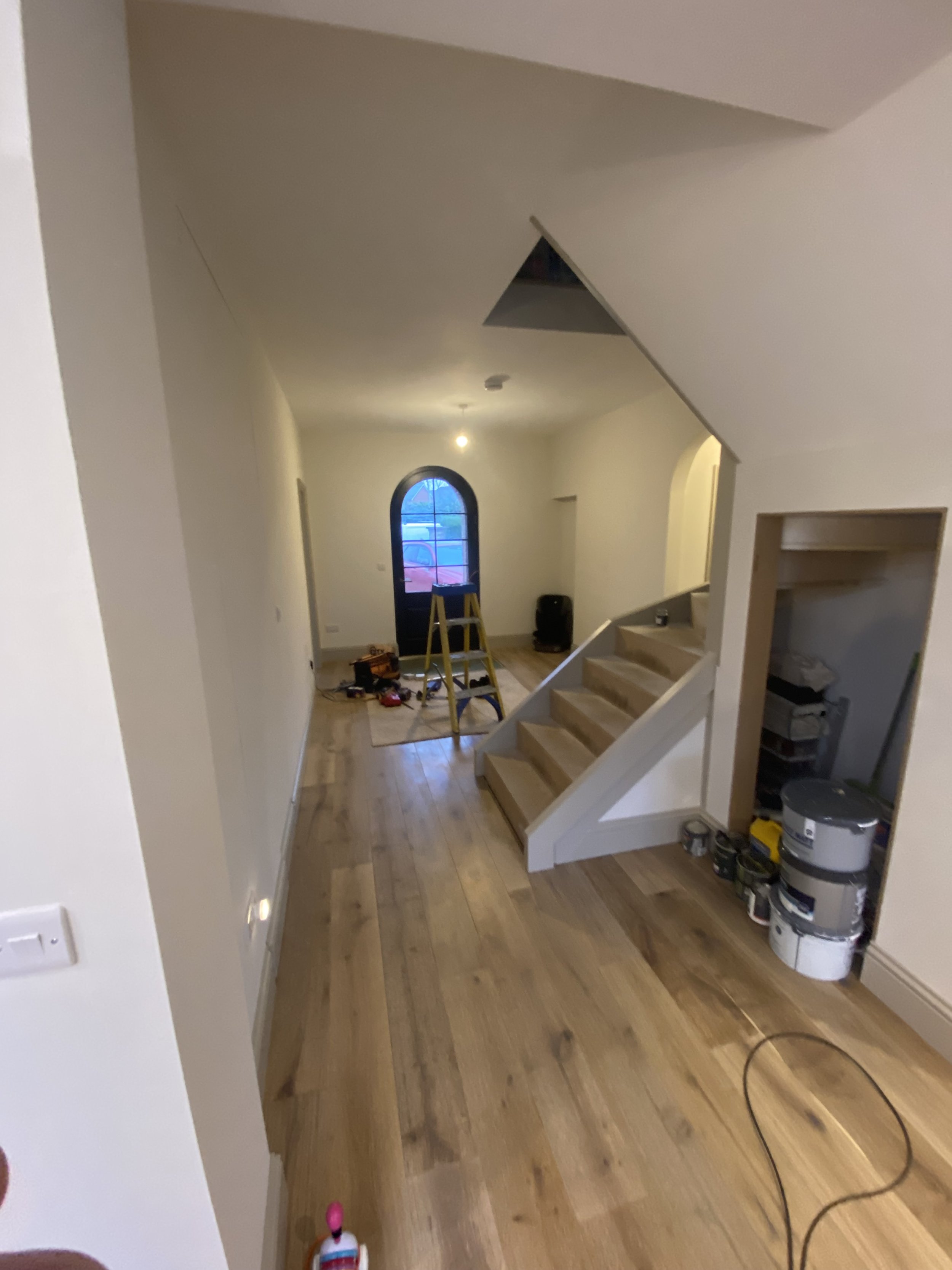 Interior of a house under renovation showing a staircase, a ladder, painting supplies, and a window with a rounded top.