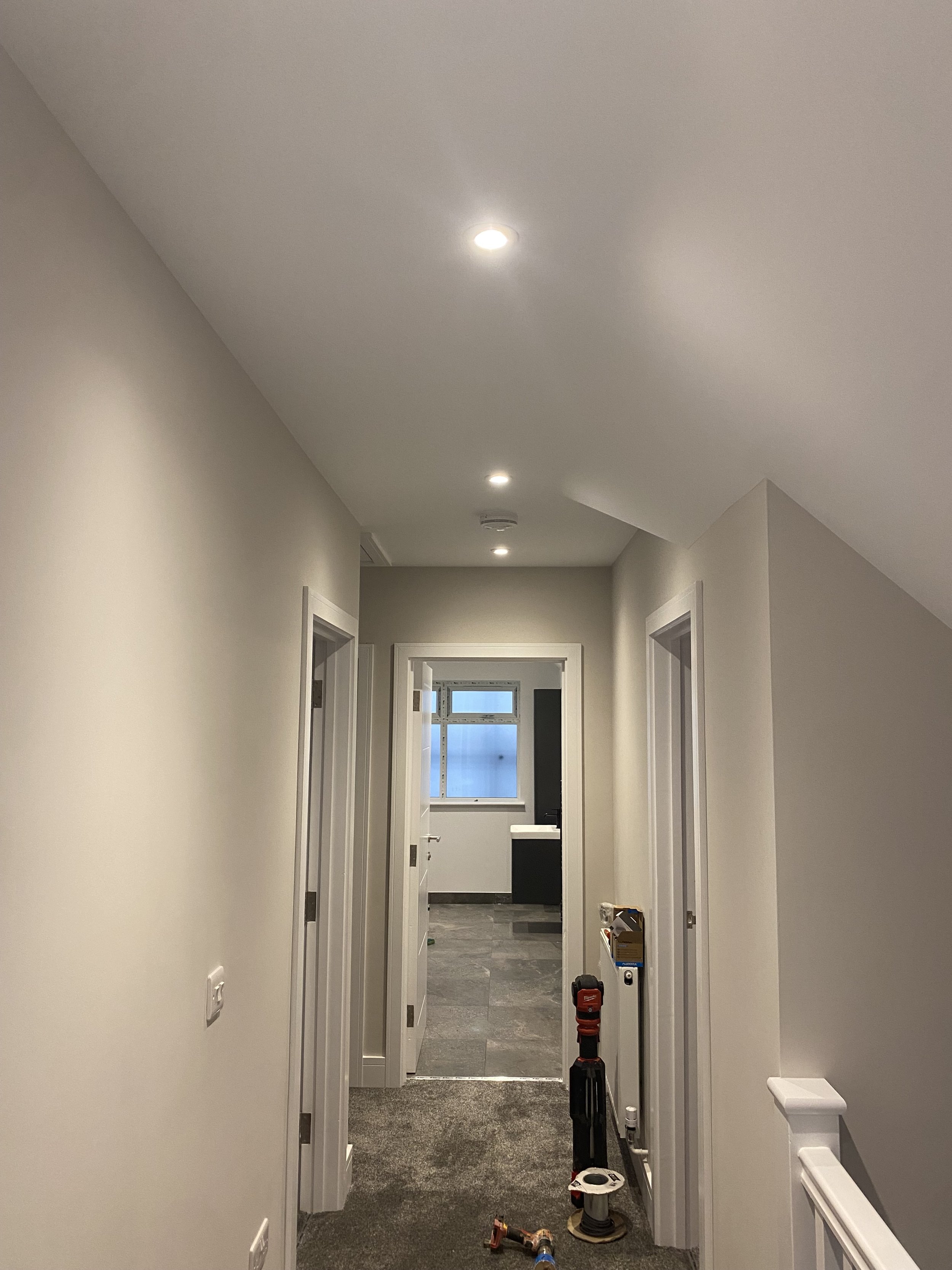 Hallway under renovation with tools and equipment, including a cordless drill and a vent pipe, on a dark gray tiled floor, beige walls, and ceiling with lights.