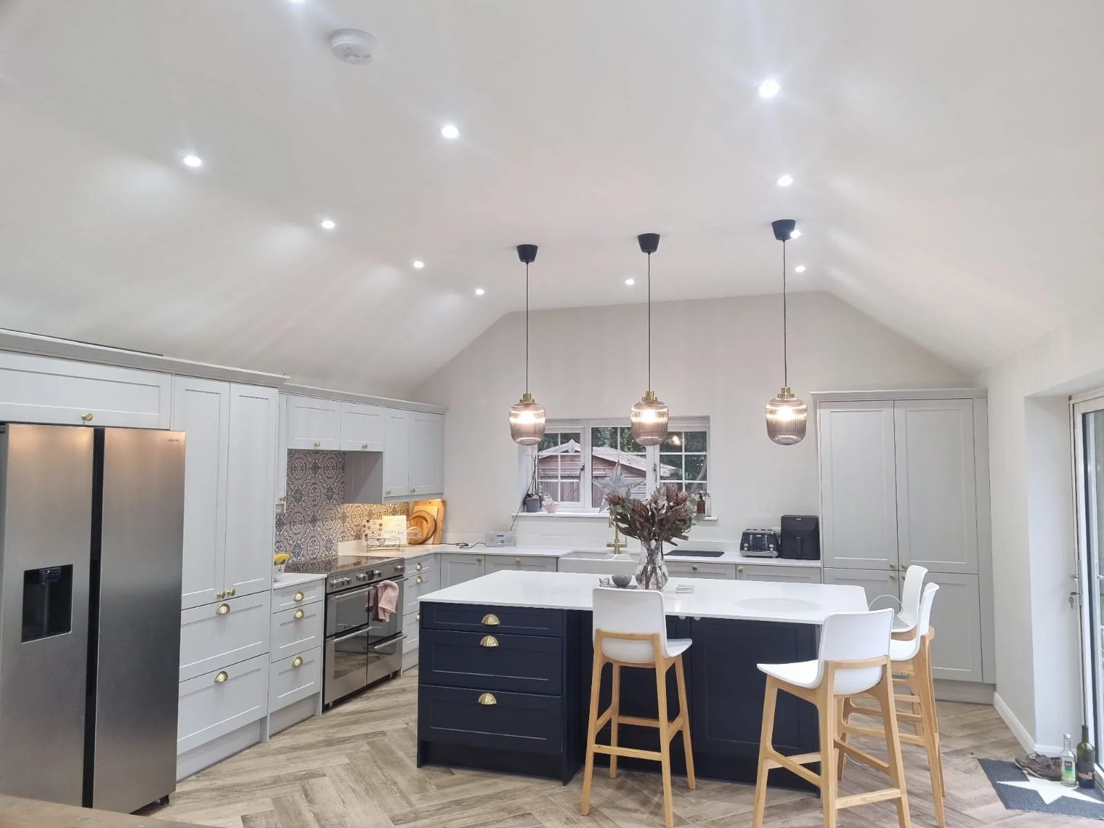 Modern kitchen with white cabinets, a navy island, and three pendant lights hanging above it. A wooden dining table with white chairs is in the foreground. A window above the sink shows an outdoor view, and stainless steel appliances are visible on t