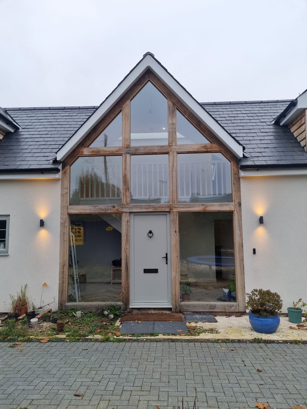 A new addition to a house under construction, featuring a large glass front with wood framing, a peaked roof with black shingles, exterior lights, and potted plants outside.