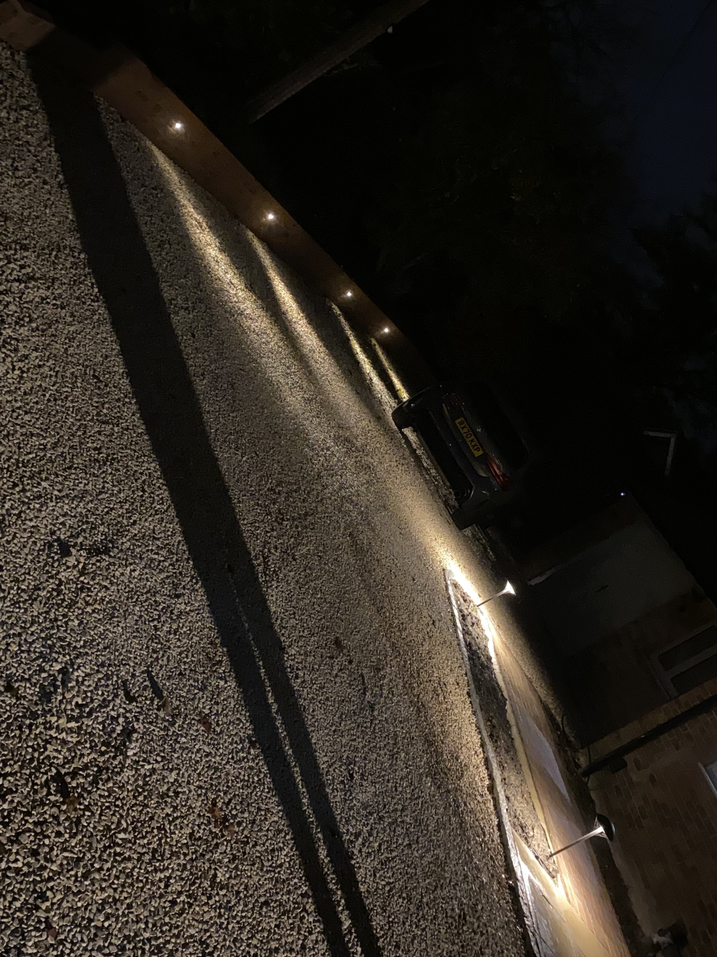 Nighttime view of a sidewalk with gravel surface, illuminated by pathway lights. Cars are parked along the street, and residential houses are visible in the background.