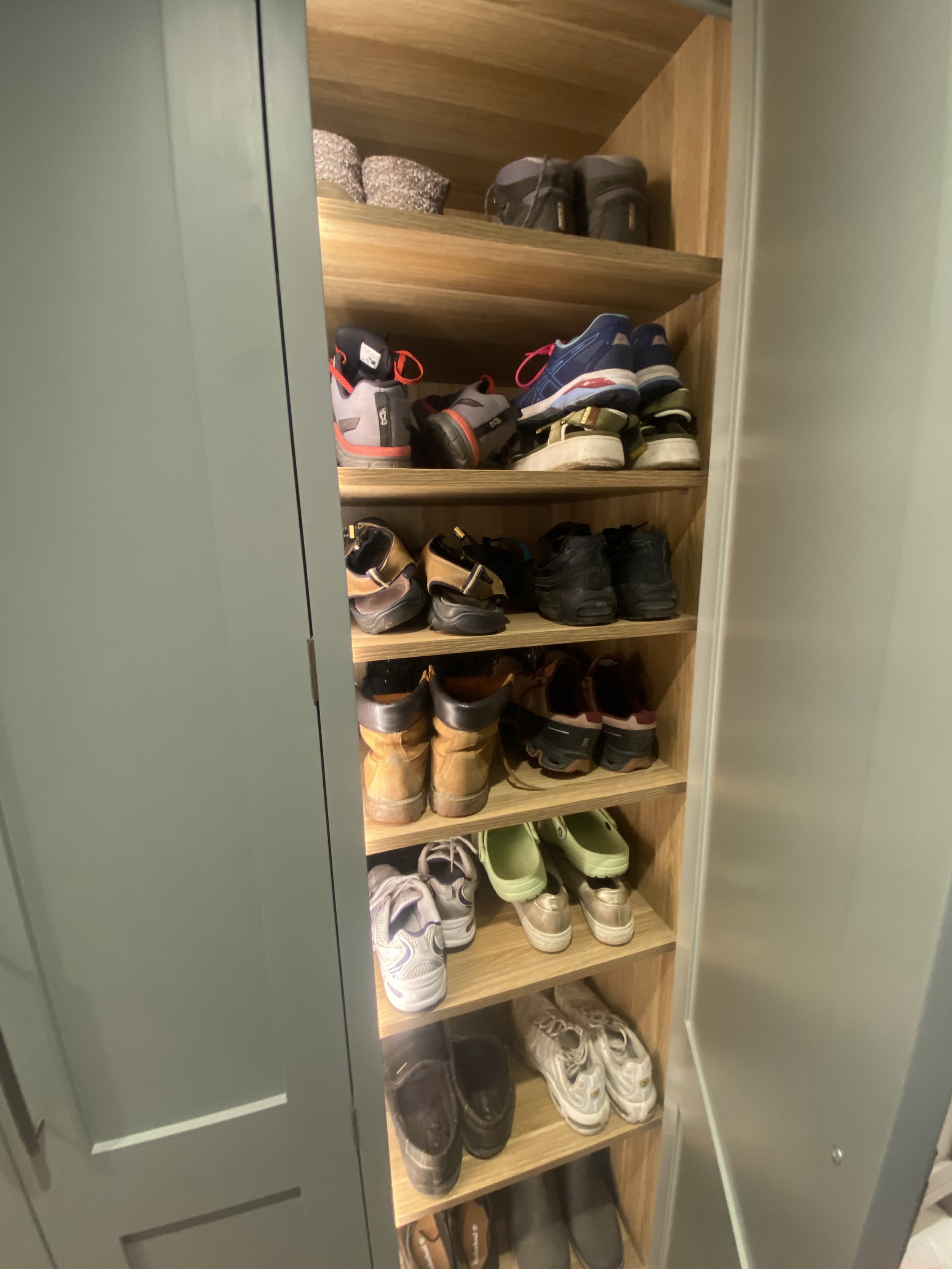 A wooden shoe rack inside a cabinet with various shoes arranged on the shelves, including sneakers, boots, and athletic shoes.