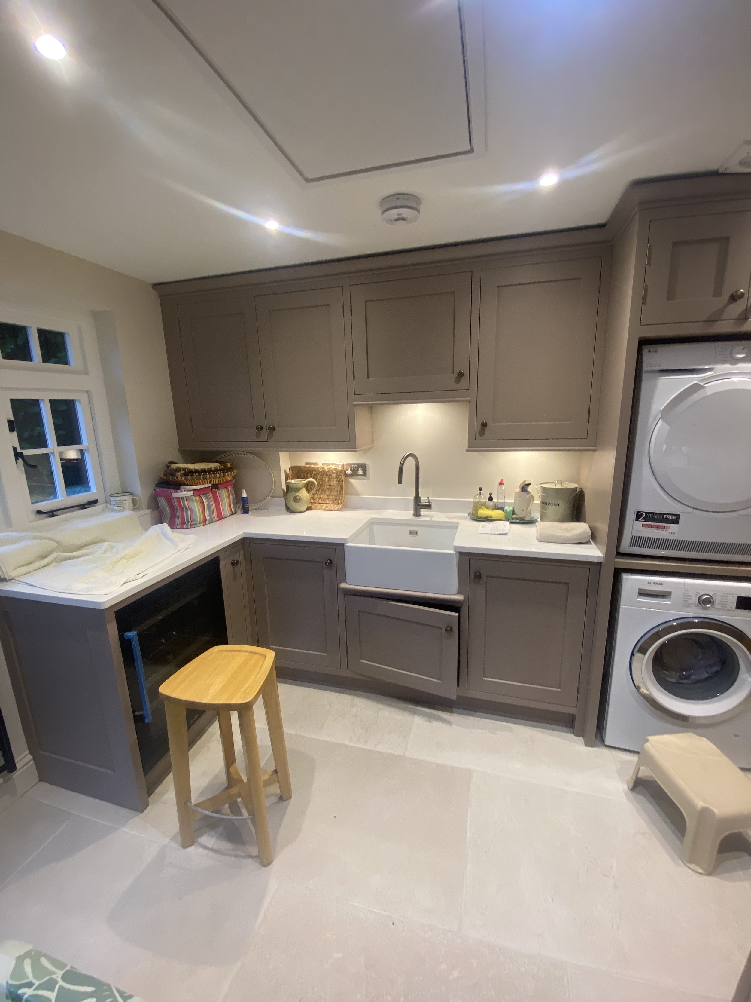 A kitchen with beige cabinets, white countertop, white farmhouse sink, and white appliances including a washer and dryer. There is a window with small panes on the left, various baskets and dishes on the counter, a wooden stool, and a small bench on 