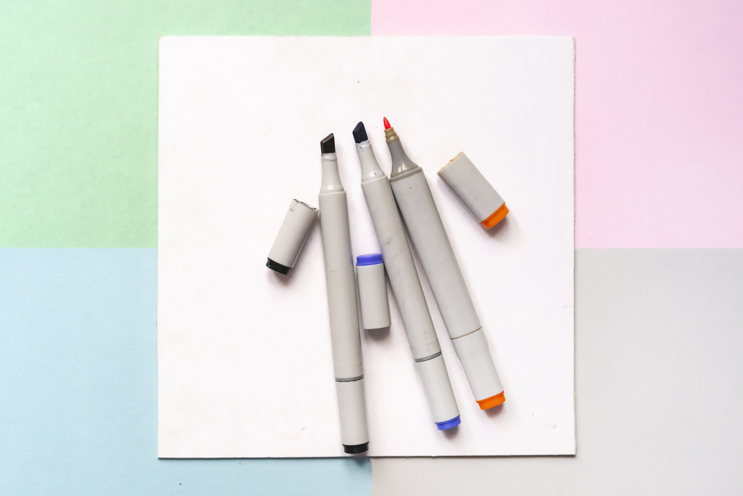 Five dual-tip markers with caps, featuring black, gray, purple, red, and orange ink, are arranged diagonally on a white sheet against a pastel-colored background with sections of green, pink, blue, and white.
