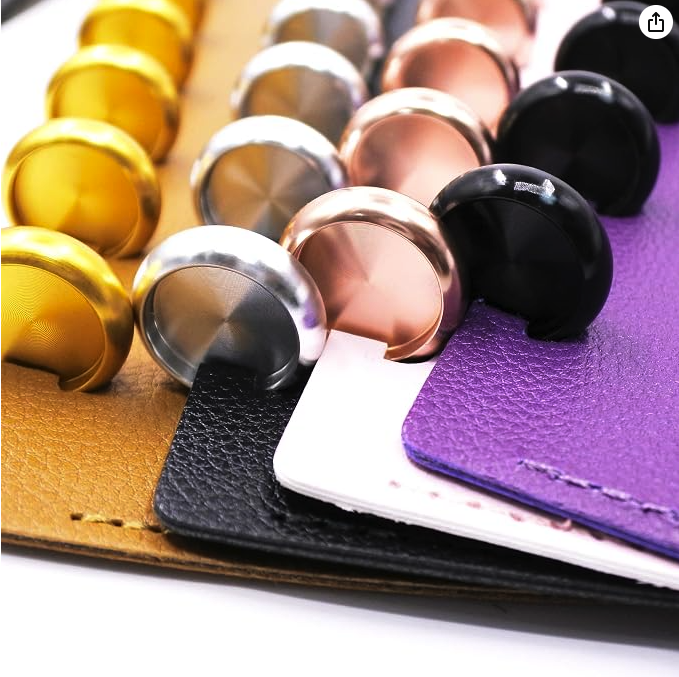 A collection of colored curtain or cabinet knobs arranged on textured faux leather and notebook covers, including gold, silver, black, and rose gold, with purple, white, and black covers.