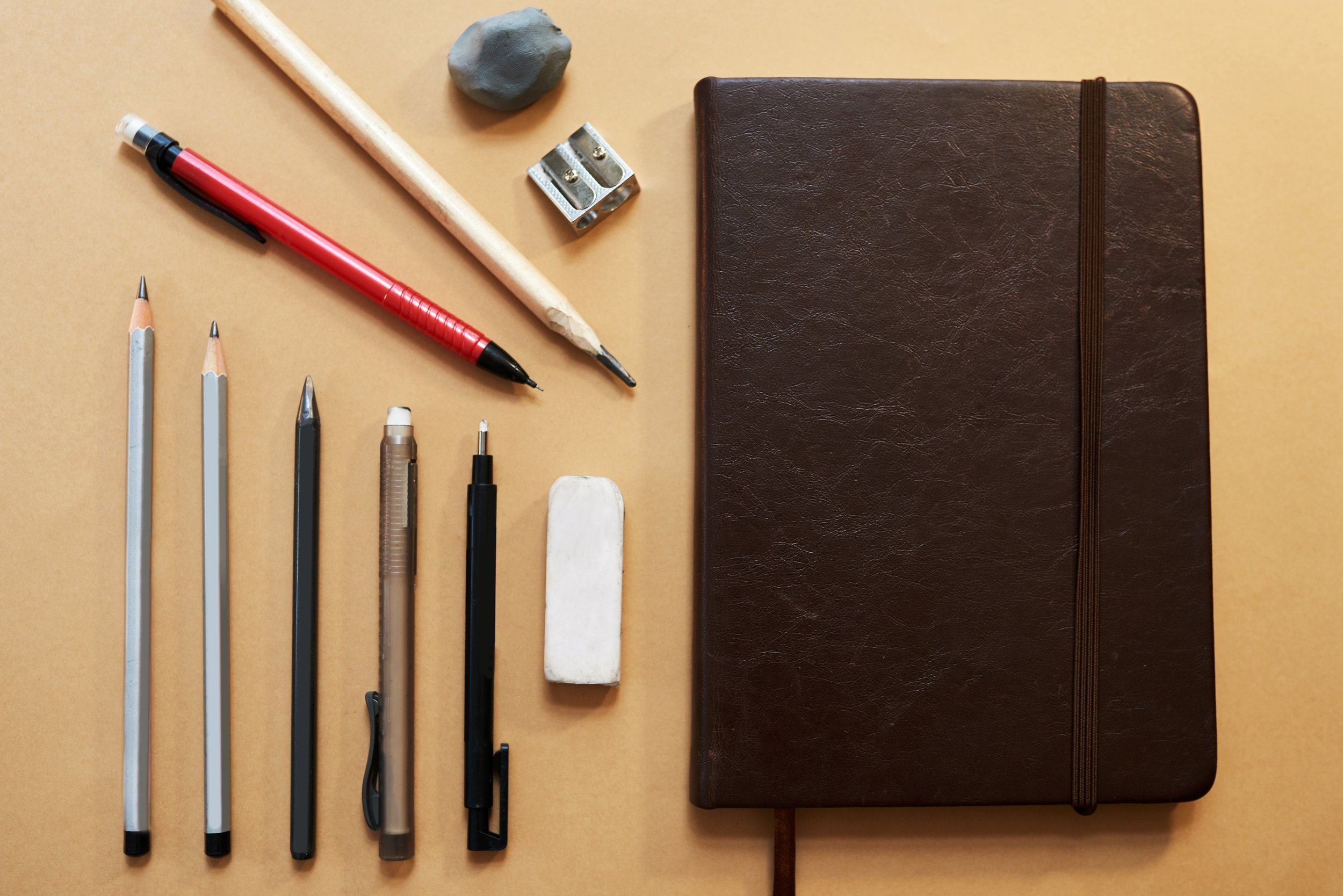 A collection of stationery items on a beige surface, including pencils, pens, a white eraser, scissors, a pencil sharpener, a piece of clay, a wooden stick, and a brown leather-bound notebook.