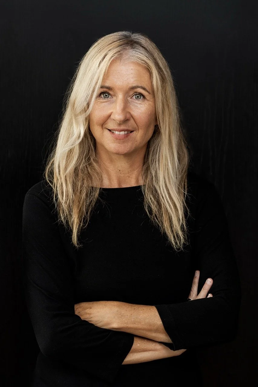 Portrait of a woman with long blonde hair, wearing a black top, standing against a dark background, smiling with arms crossed.