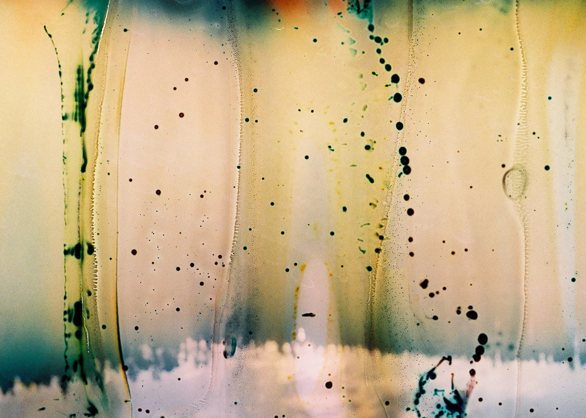 Abstract artwork featuring splashes of black, green, and yellow ink on a light background with vertical streaks and droplets.