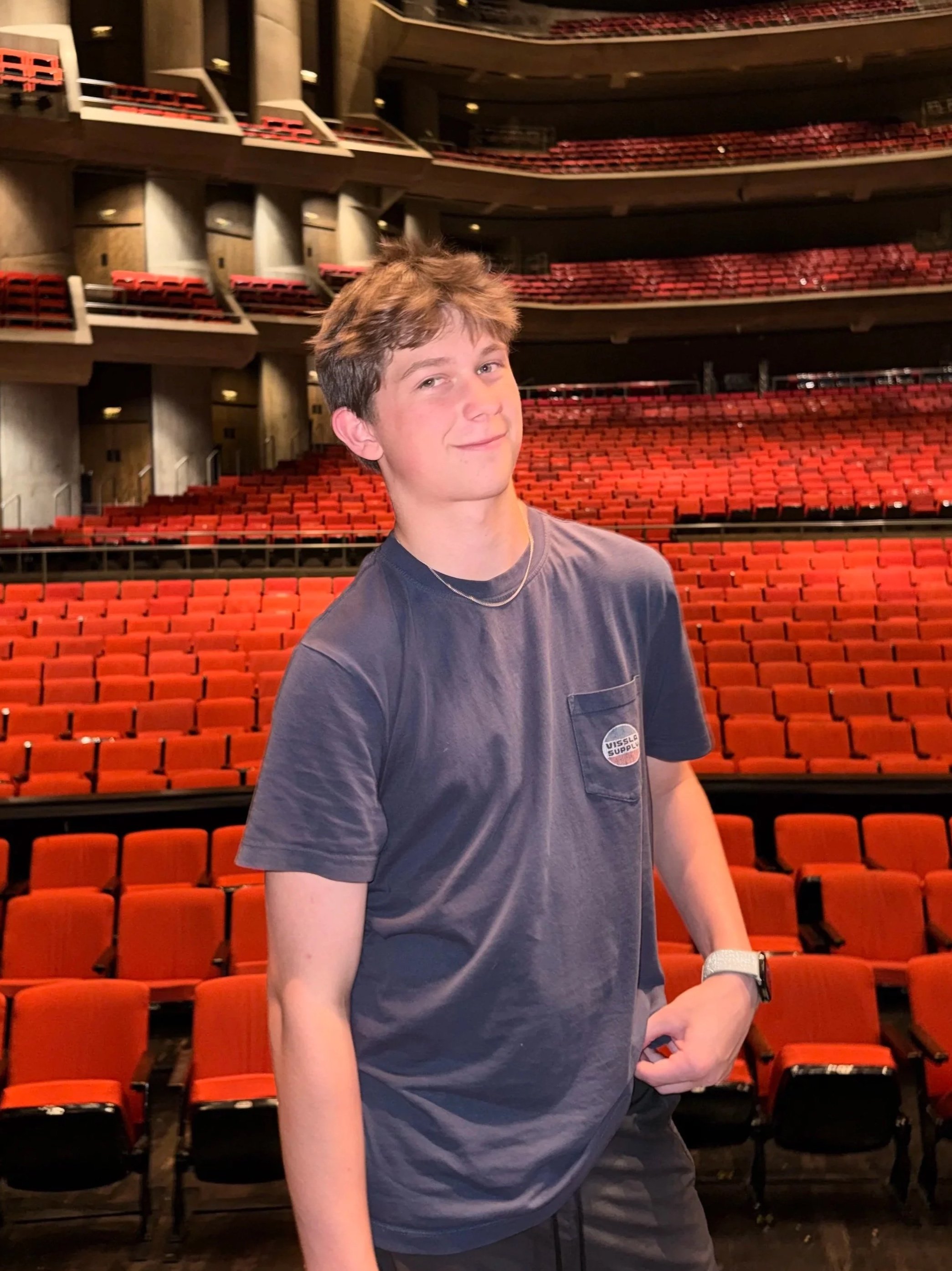 Noah Chandler with light brown hair and fair skin standing in an auditorium with red theater seats. He is wearing a dark grey t-shirt, a watch, and a necklace, and has a slight smile on his face, filmmaker, director, and actor.