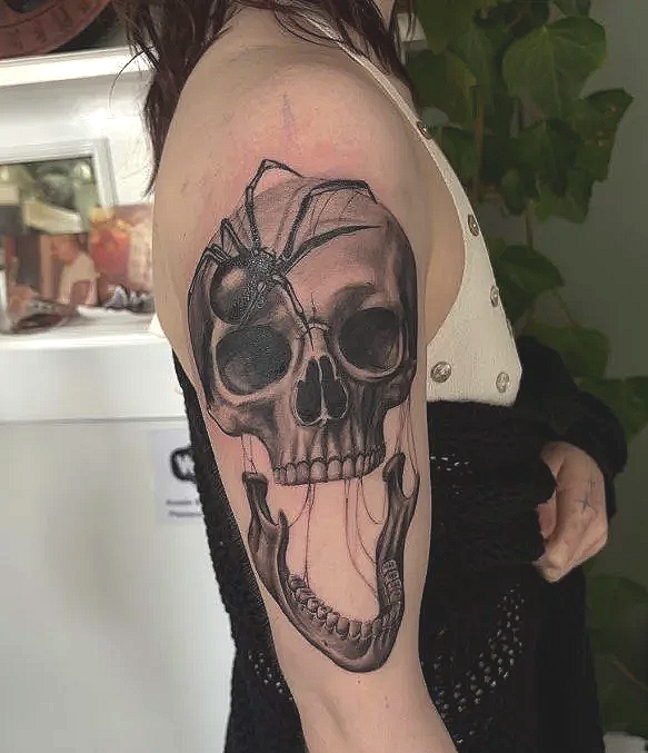 Tattoo of a skull with a spider on top and long strings hanging, in black and gray ink, on a person's upper arm.