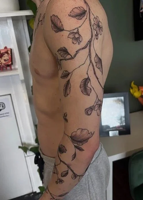 A person's side view with a black outline tattoo of a leafy vine running from the shoulder down the arm.