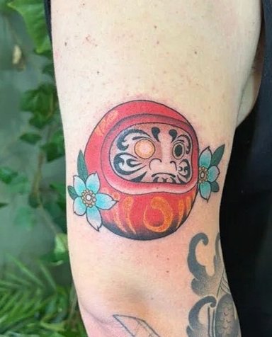 Tattoo of a Daruma doll with floral accents on a person's arm.