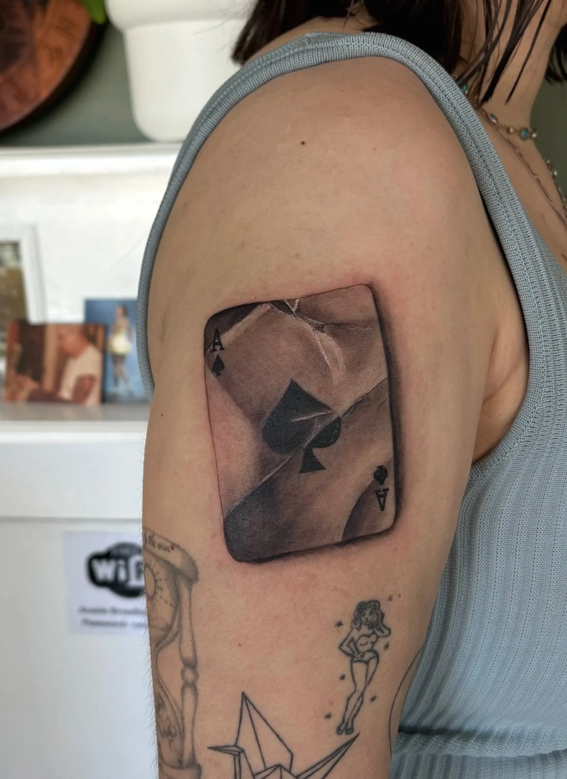 A person's upper arm with multiple tattoos, including a large playing card with a spade symbol, a pin-up style girl, a paper airplane, and a fairy. The arm is partially covered by a sleeveless shirt, and the background features a blurred picture and 