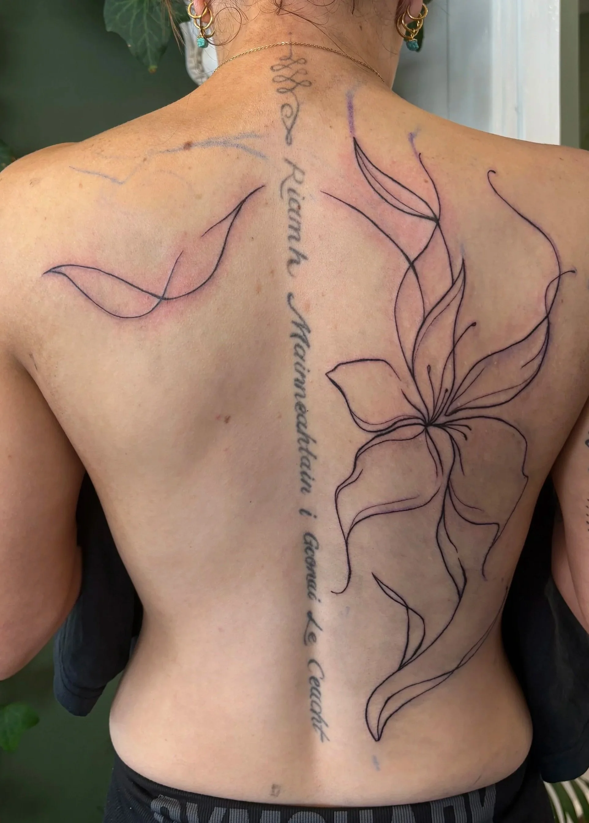 Back view of a person with a large outline of a flower tattoo on their back, accompanied by cursive text running down the center of their back. The person is wearing earrings and a delicate necklace, with short hair and a skin tone that is light to m