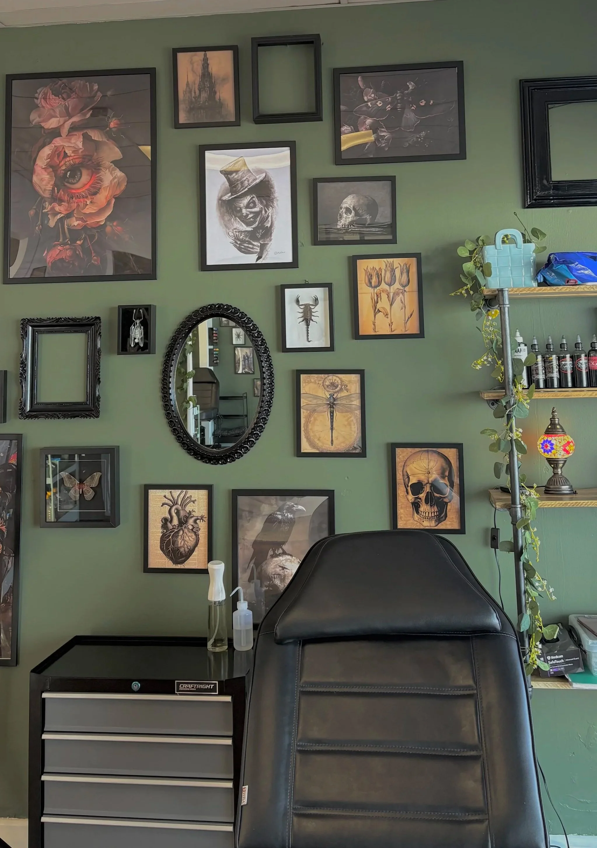 A wall decorated with various framed artworks including an eye with flowers, a castle, a skeleton, a scorpion, a dragonfly, and other artistic images. A mirror is also hanging on the wall, and there is a shelf with plants and bottles. In the foreground, there's a black leather chair, a small black and gray storage cabinet, and bottles of hand sanitizer.