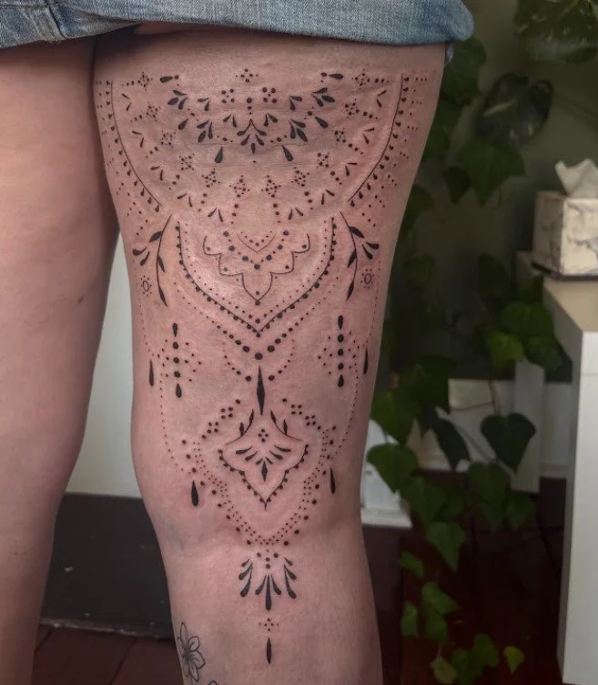 A tattoo on a person's thigh featuring symmetrical black linework and dot patterns in a decorative, mandala-inspired design.