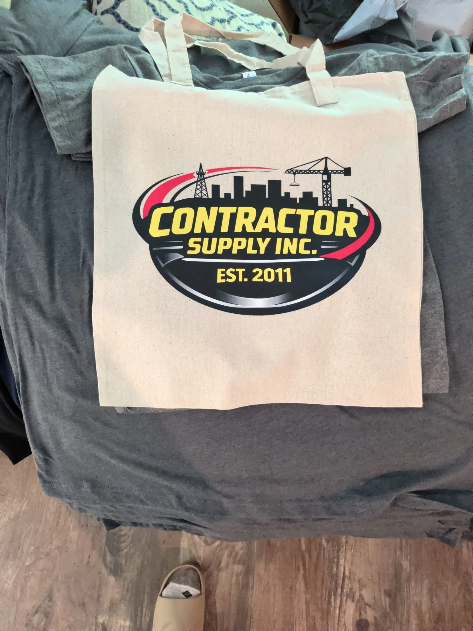 A paper tote bag with a logo for a construction supply company, placed on a crumpled gray tablecloth with a person's foot visible at the bottom of the image. The logo features a city skyline, a crane, and an oil rig, with the text "Contractor Supply Inc." and "Est. 2011."