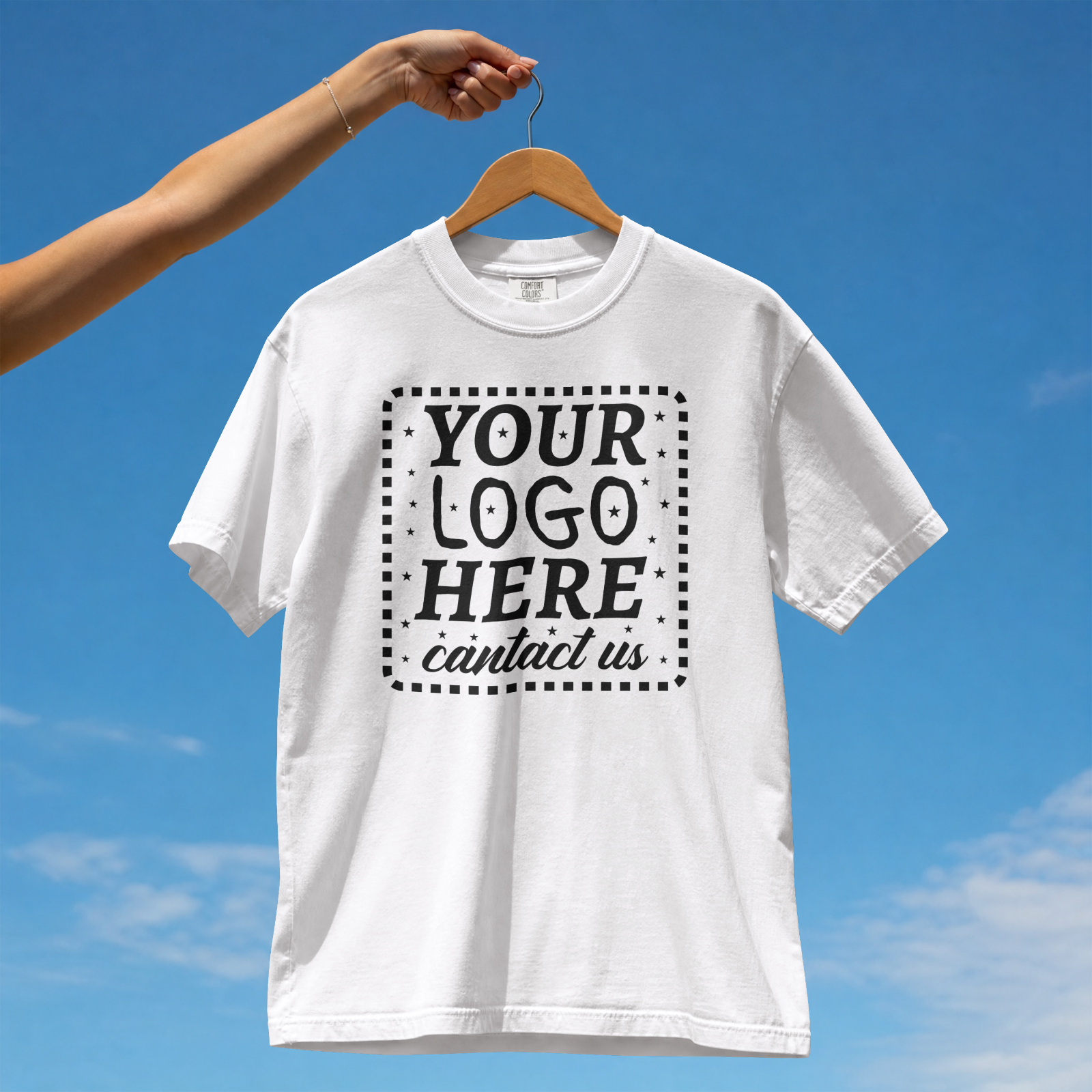 A white T-shirt hanging on a wooden hanger being held up against a blue sky background. The T-shirt has placeholder text that says: "YOUR LOGO HERE contact us" surrounded by small stars and dots.