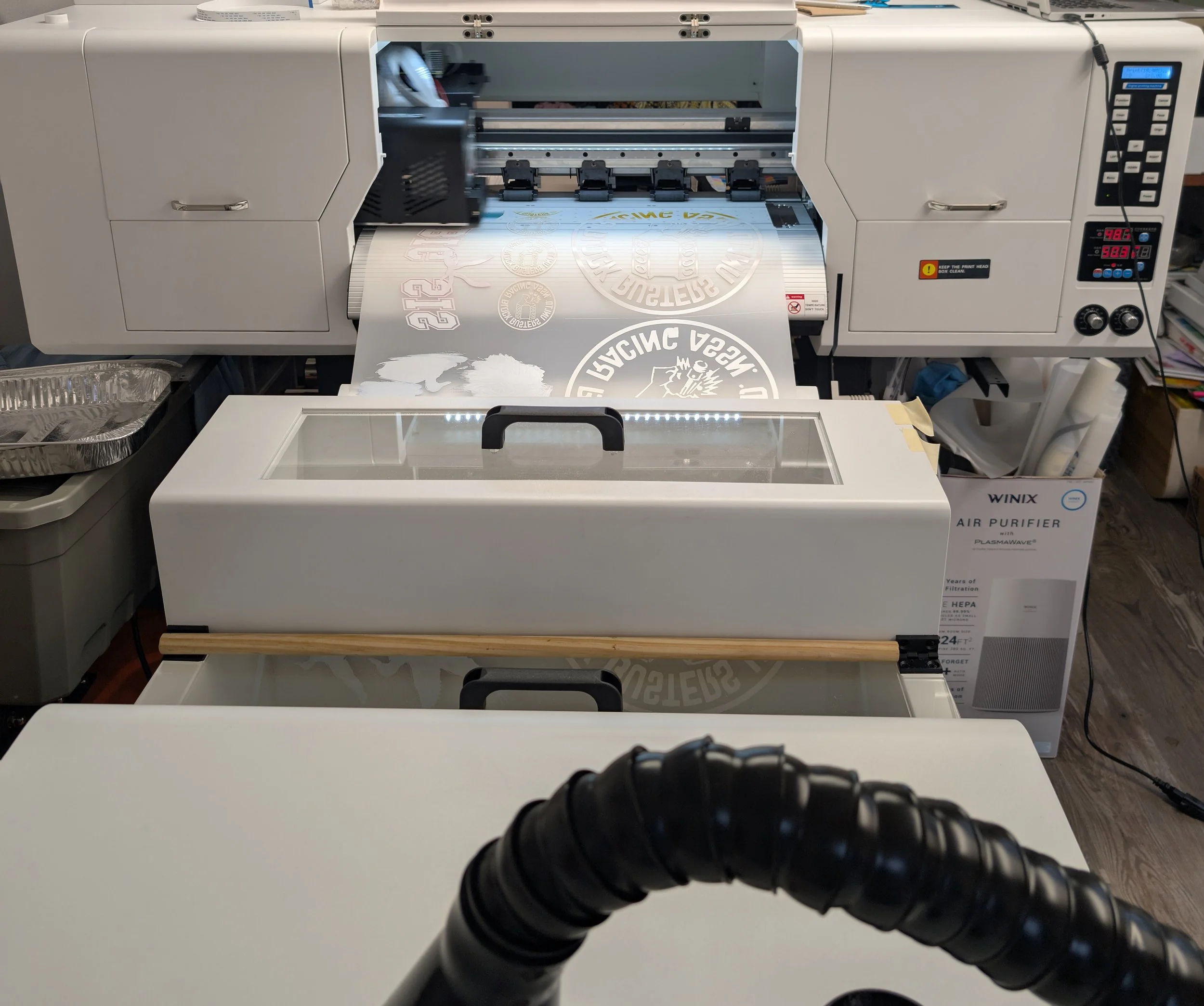 A DFT Printer printing a gangsheet of images. Some are large round graphics and some smaller. The images are going into the curing oven to be powdered and cured.