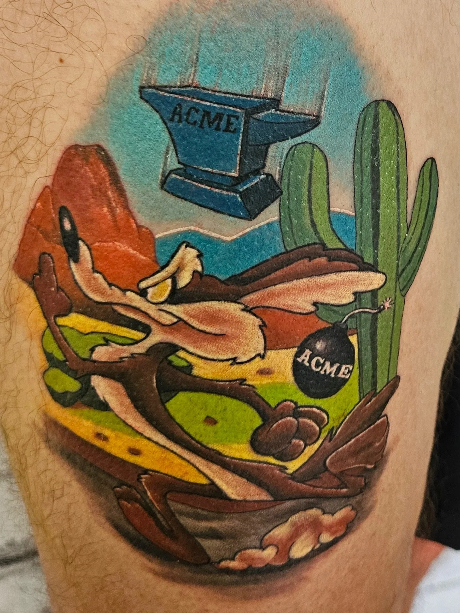 Colorful tattoo depicting a desert scene with cacti, mountains, a yellow road, a black bomb with 'ACME' written on it, and a dynamite stick, in a cartoon style. Also a scar cover tattoo.