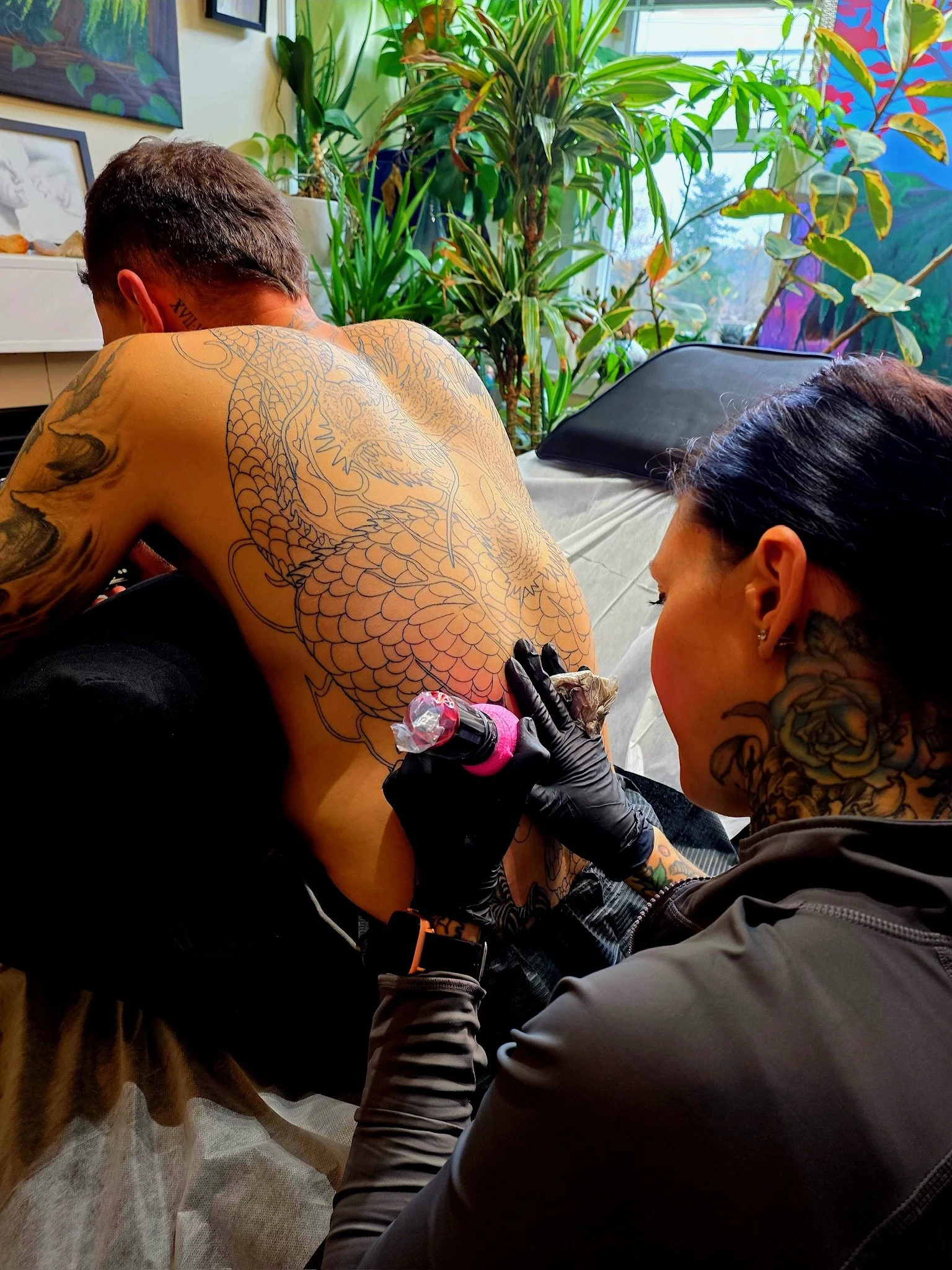 A person with tattoos, presumably getting a tattoo, with another person working on the tattoo, both wearing black gloves. They are in a room with large green plants and artwork in the background.