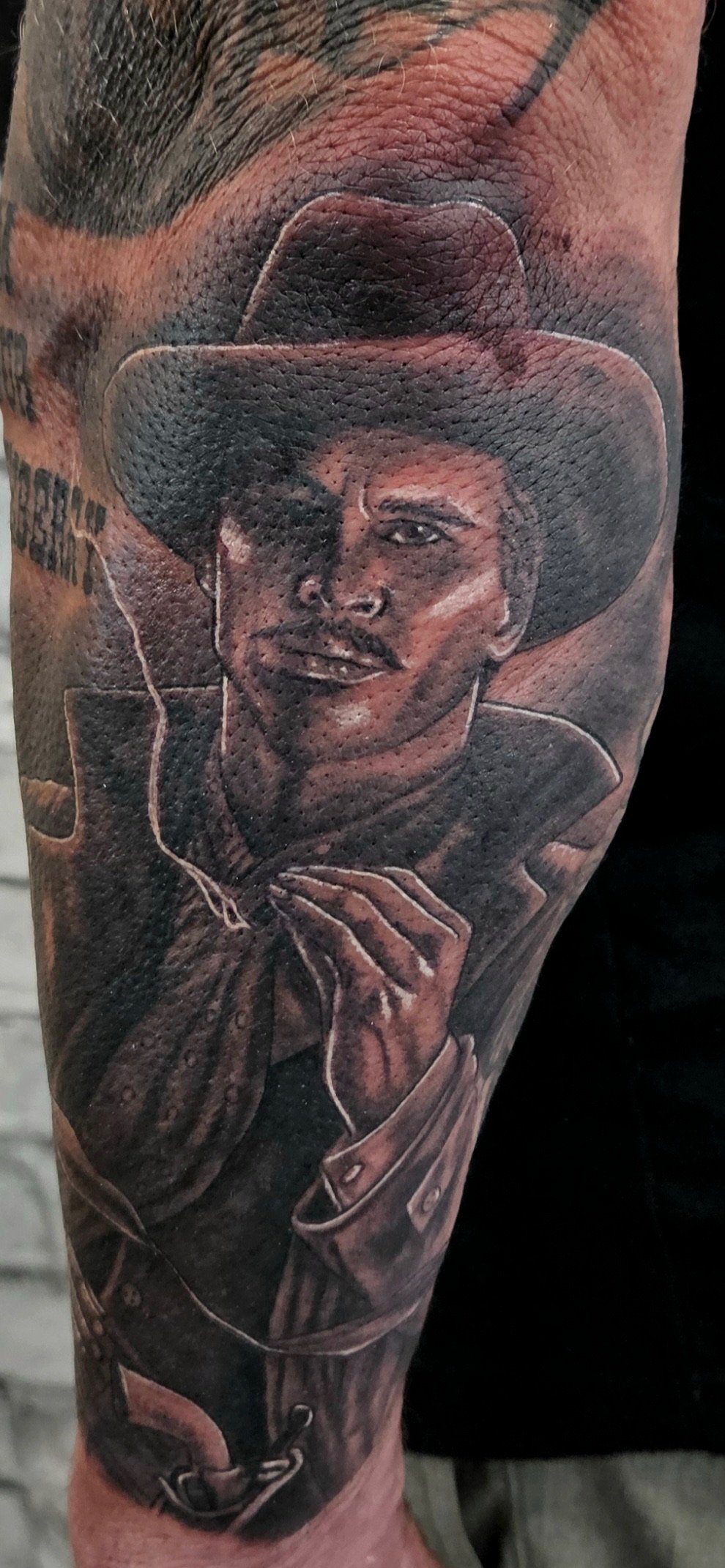 Black and gray tattoo of a cowboy wearing a wide-brimmed hat, with a confident smile, on a person's arm. Black and grey realism.