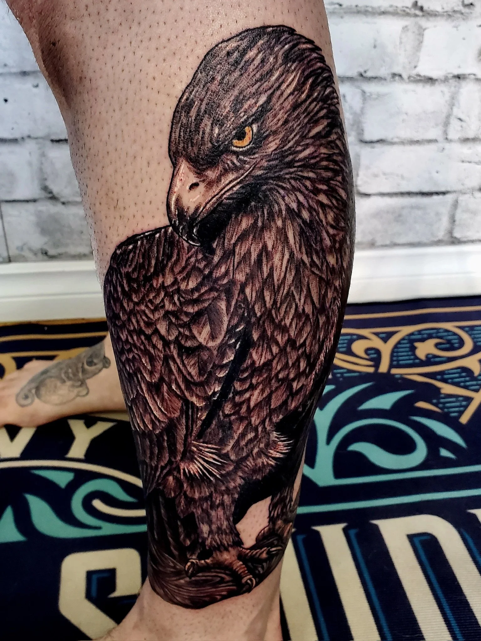 Tattoo of a black eagle's head with yellow eyes on a person's leg, with a brick wall in the background. Black and grey realism.