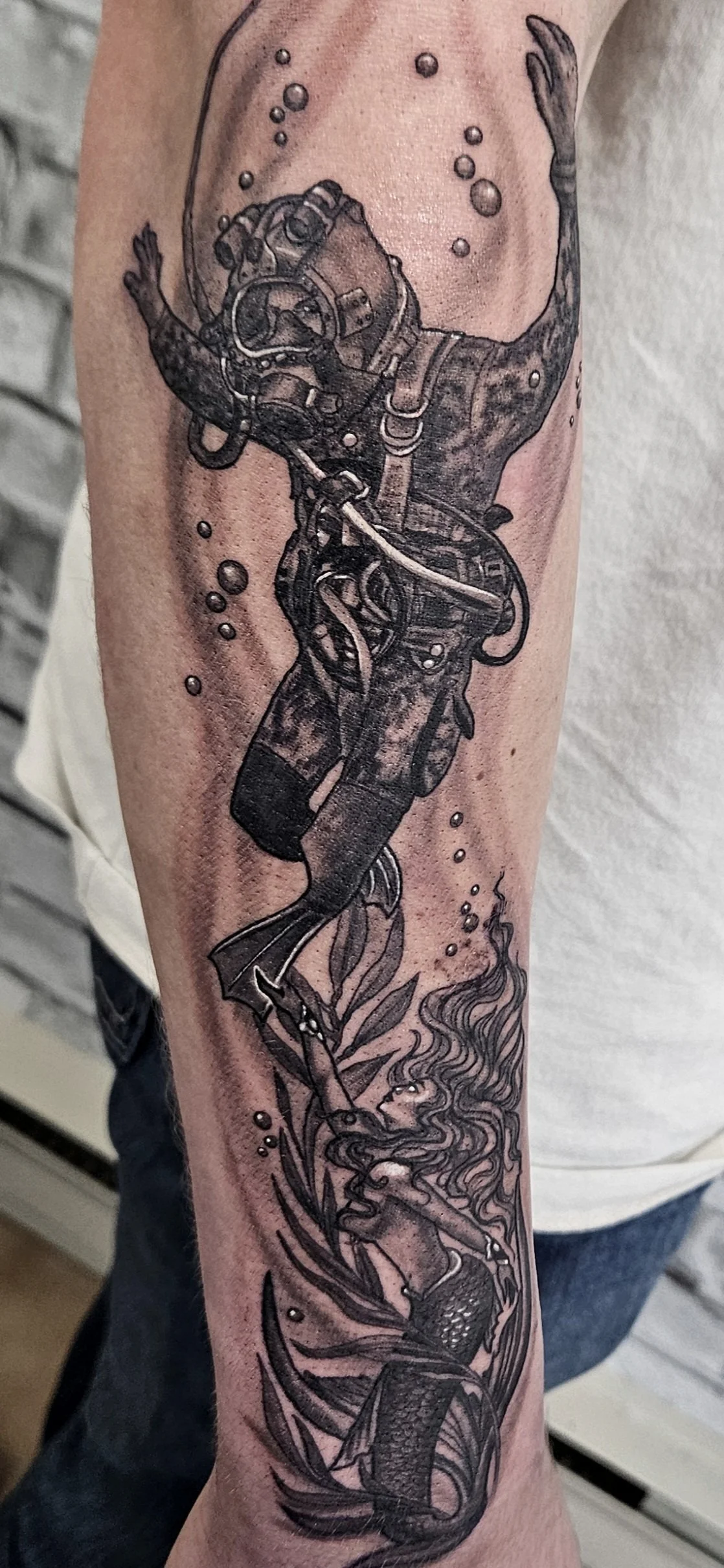 Tattoo of a deep-sea diver with a tank, rising above a mermaid with flowing hair ocean plants, and bubbles around.