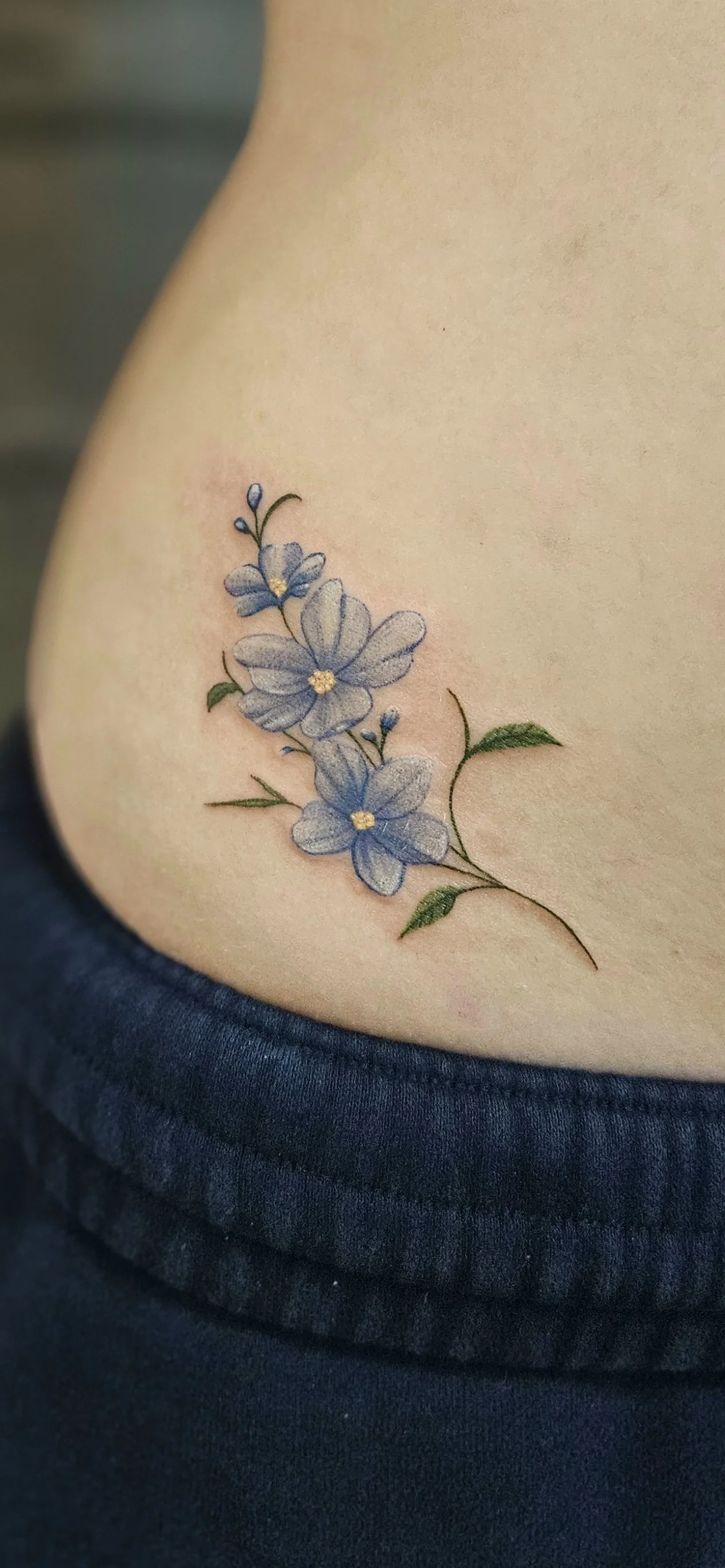 Close-up of a tattoo of blue flowers with green leaves on a person's hip. Fine line design, delicate floral tattoo, very feminine.