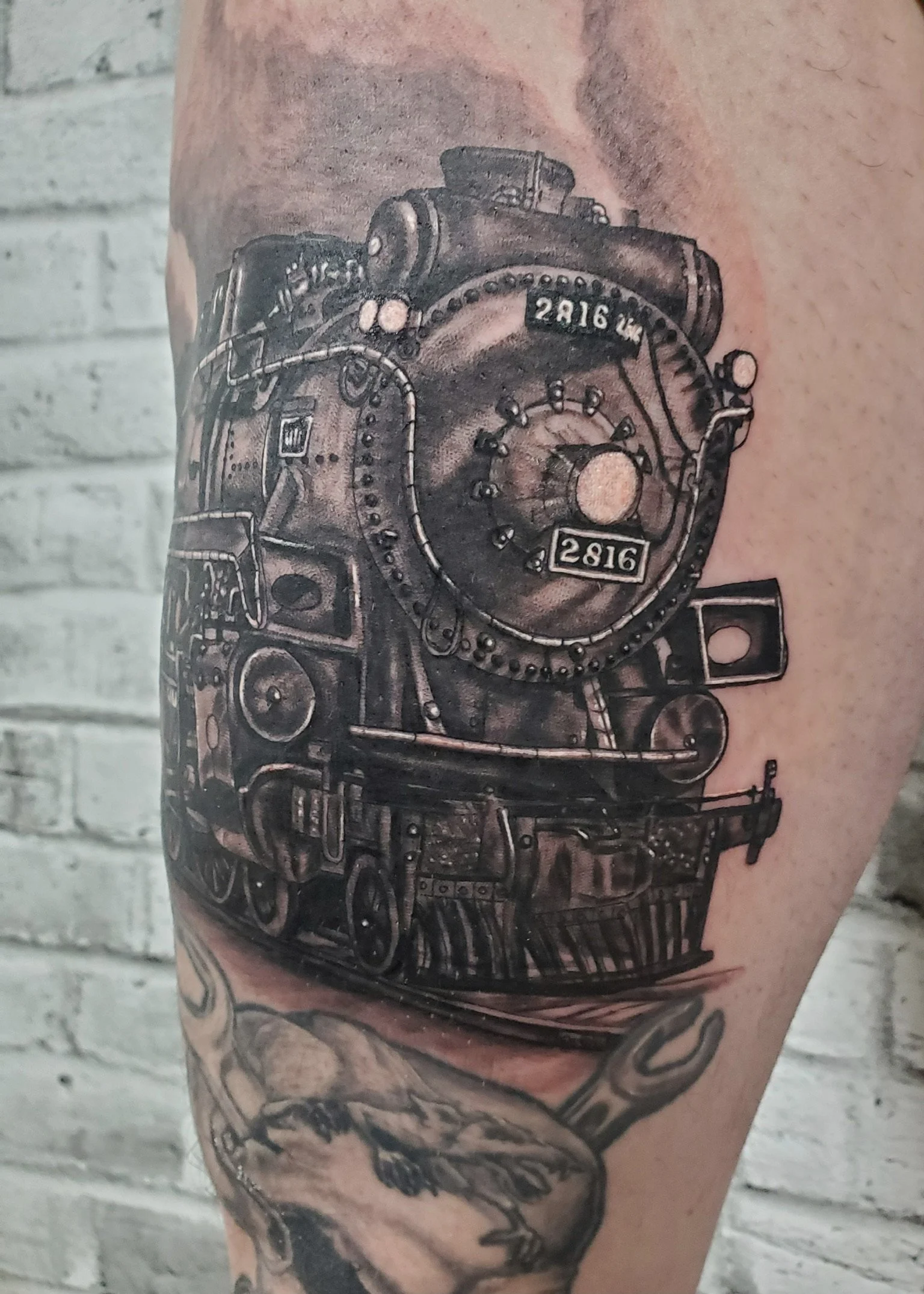 A detailed black and gray tattoo of a vintage train engine with the number 2816, covering a person's leg.