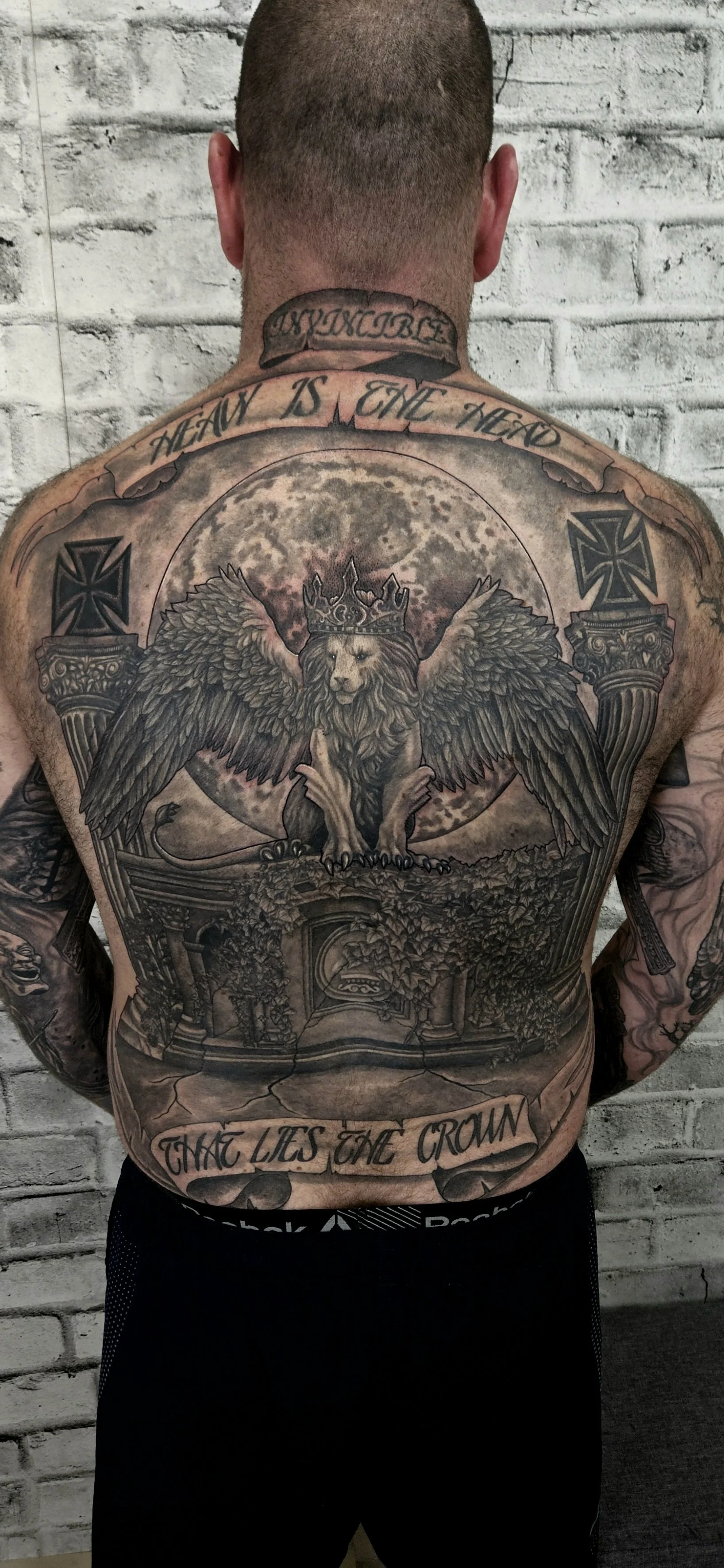 A man with a full Backpeice tattoo of a winged lion with a crown, standing on ruins with ivy, moon in the background. Black and grey realism.