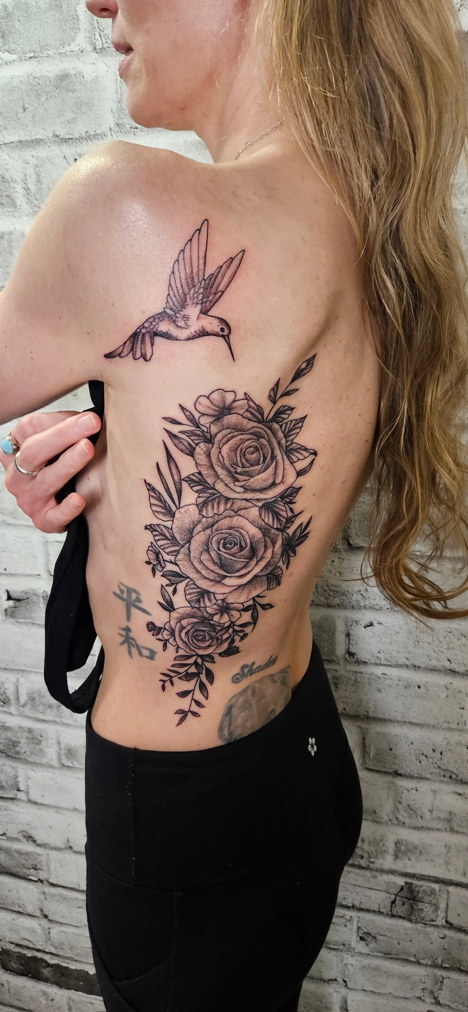 A woman with long red hair and light skin showing a large tattoo on her side and shoulder, which includes a hummingbird and three roses with leaves.