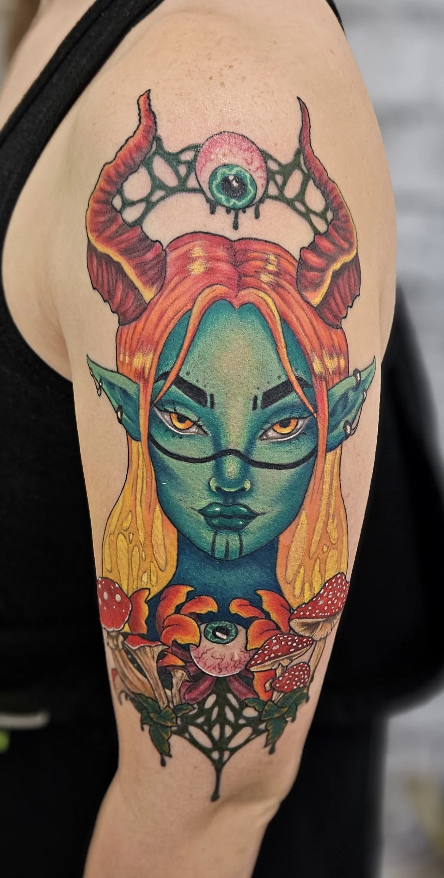 Colourful tattoo of a fantasy female elf with green skin, orange hair, and pointed ears, with a third eye on her forehead above her. The tattoo includes mushrooms, eyes, and decorative elements. Full colour tattoo by Jessica Scott.