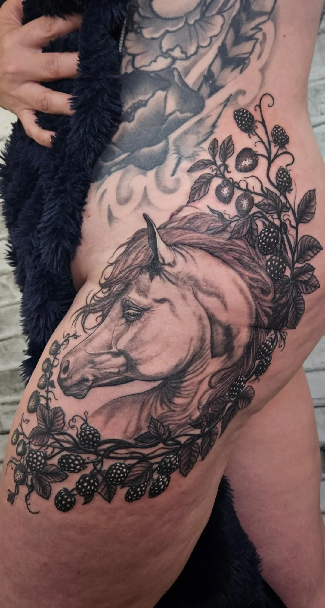 Tattoo of a horse's head surrounded by blackberry branches and leaves on a person's hip and leg. Black and gray realism.
