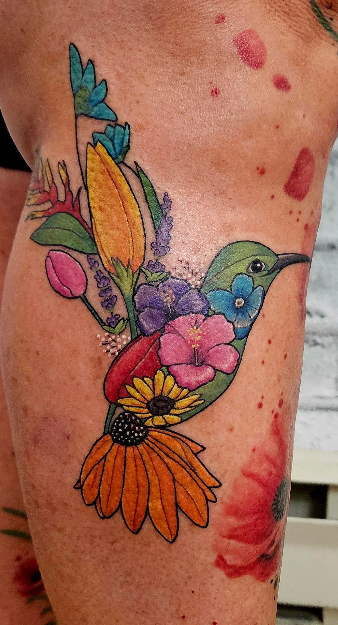 Colorful tattoo of various flowers and a hummingbird on a person's arm.