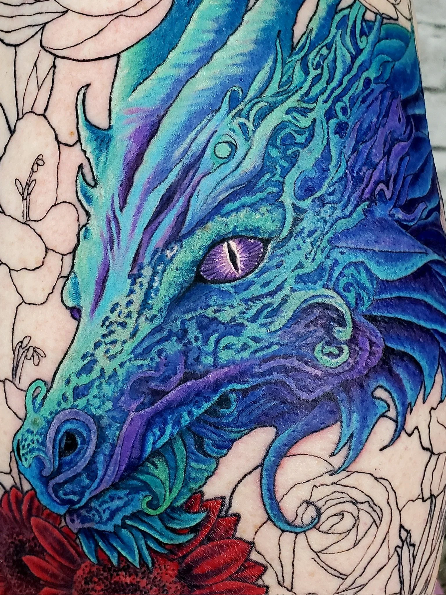 Colourful tattoo artwork featuring a dragon's face with purple eyes, surrounded by blue and green scales and intricate black outlines.