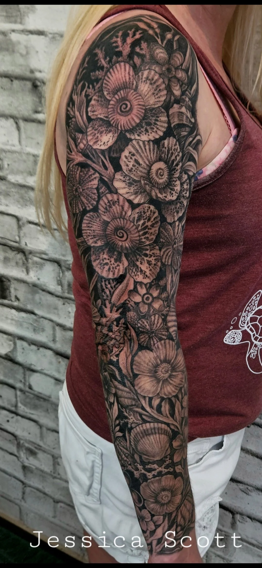 Close-up of a person's arm with a detailed black and grey floral tattoo, featuring large, intricate flowers and leaves, against a brick wall background. Ocean inspired, the flowers are sea shells and sea weed. Full sleeve realism. 