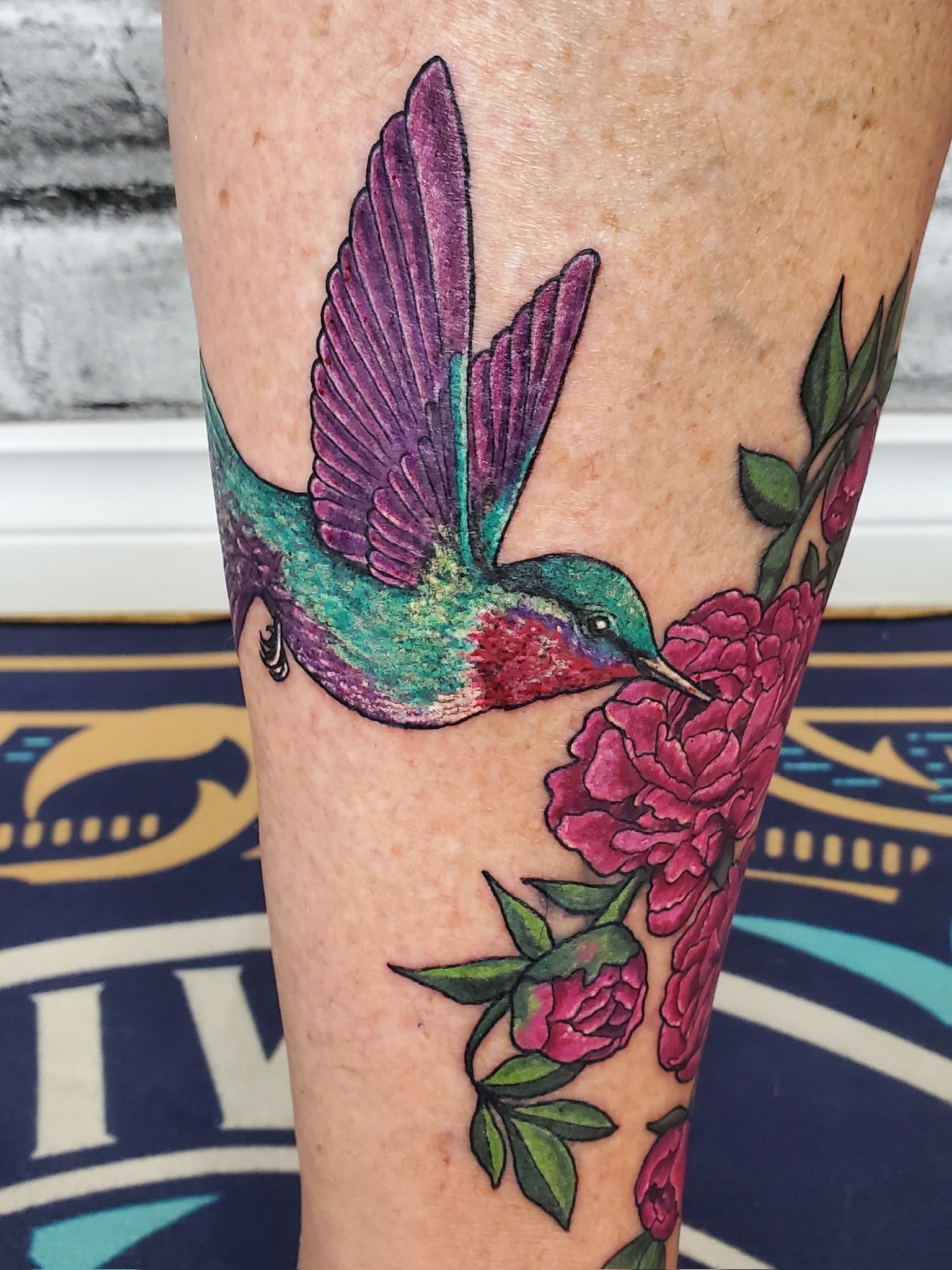 Colorful tattoo of a hummingbird with purple and green feathers perched on pink flowers with green leaves on a person's arm.