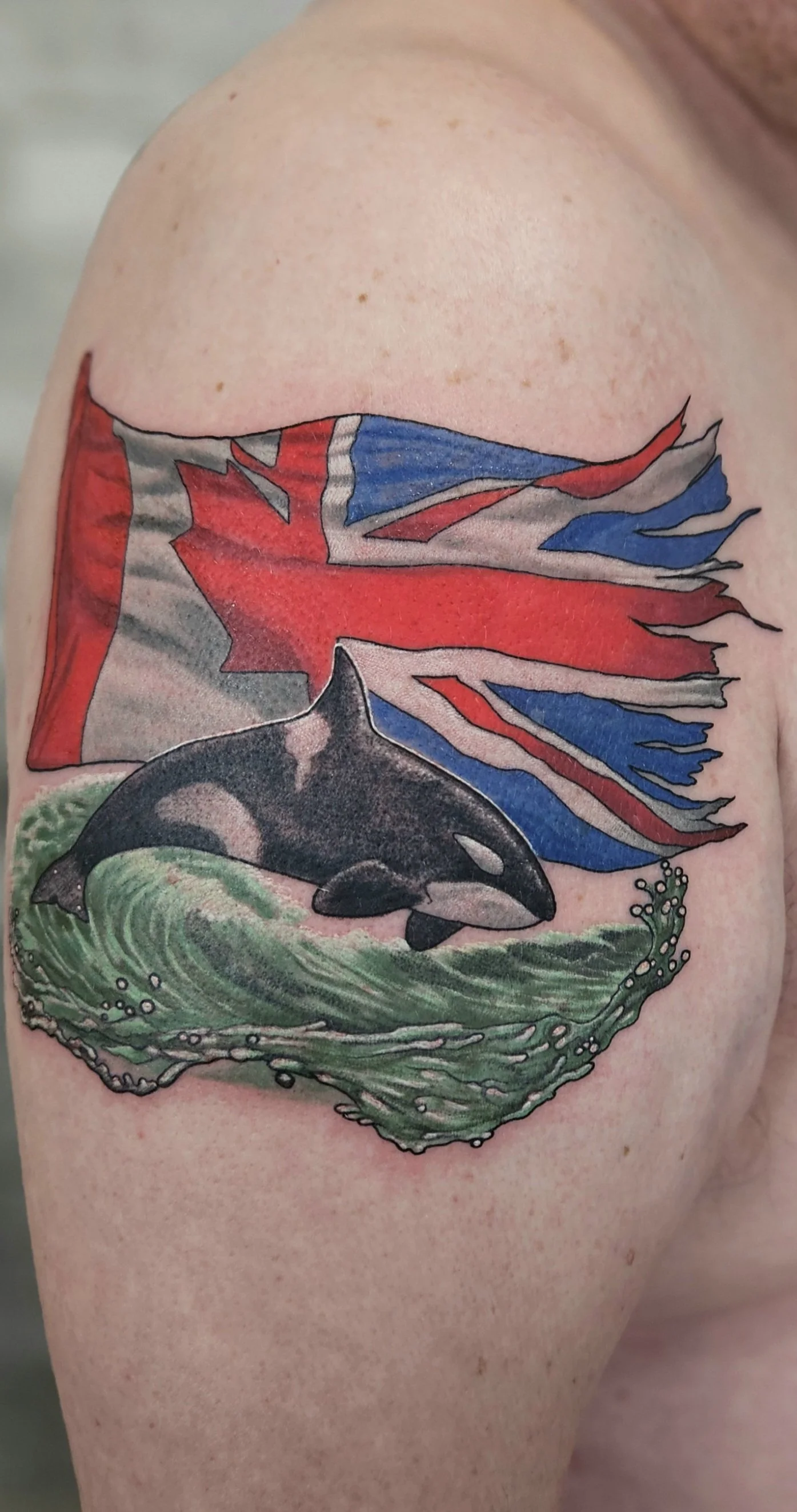 Tattoo of a whale jumping out of waves beneath a torn flag on a person's upper arm. Colour and realism. 