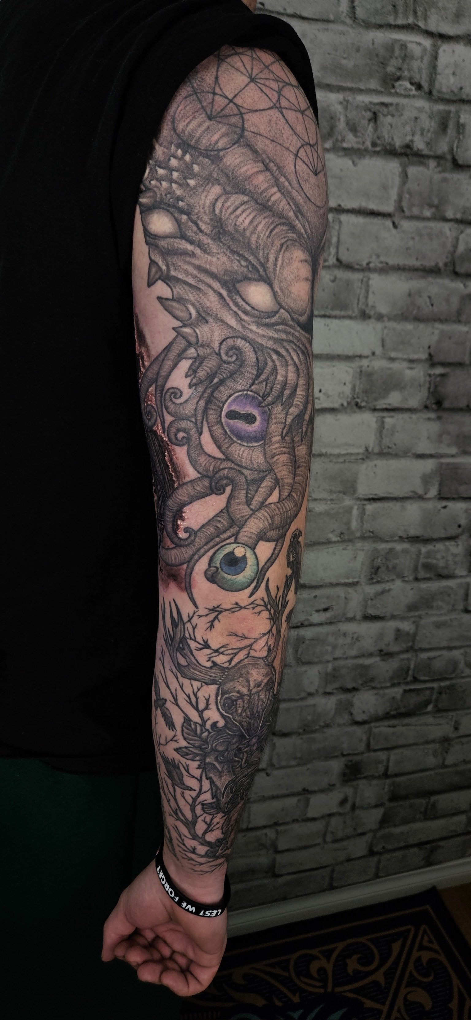 Tattoo sleeve on person's arm featuring a dragon, eyes, tree branches, and abstract designs, in black and gray ink with some color accents.