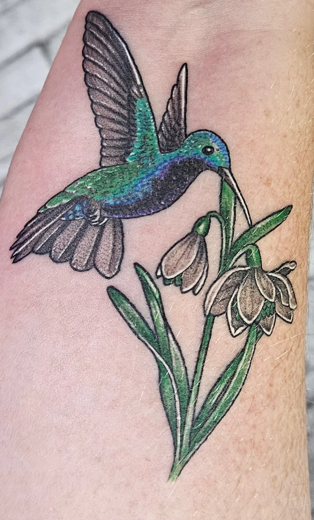 Tattoo of a colorful hummingbird and flowers on skin.