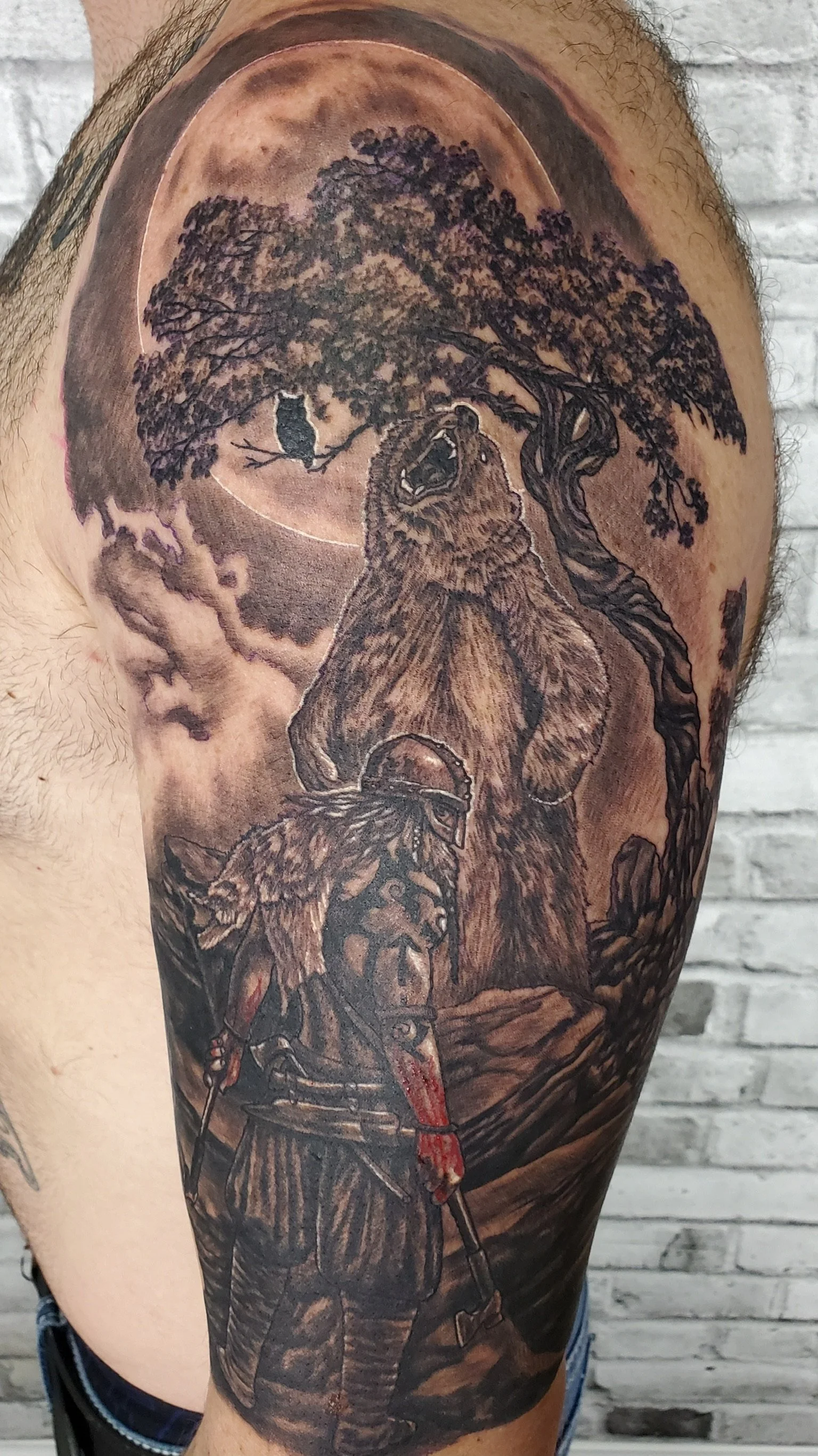 Tattoo of a grizzly bear, a tree, an owl, and a soldier with a helmet and sword under a night sky with a full moon. Black and grey realism on person upper arm.