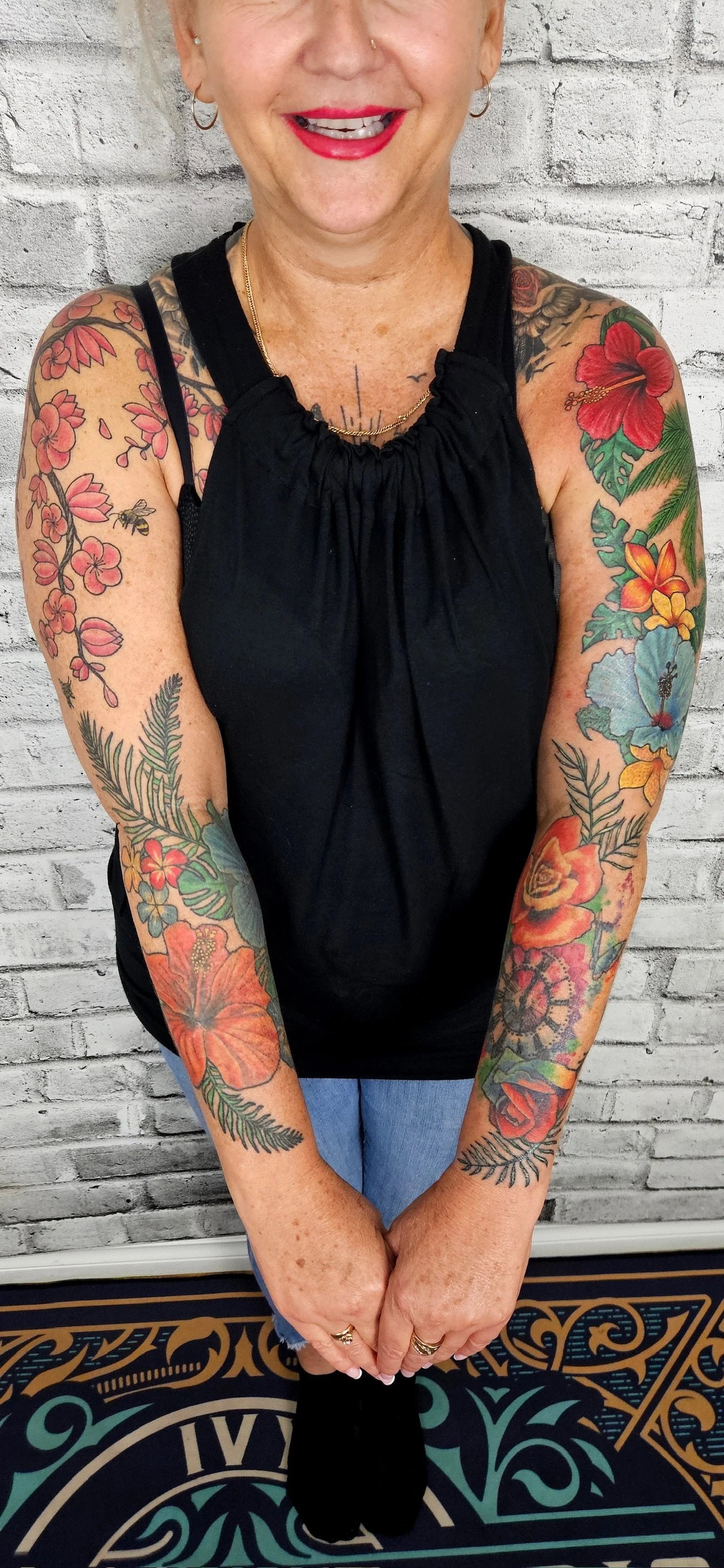 A woman with short blond hair, wearing a black sleeveless top, smiling with red lipstick, and showing colorful floral tattoos on both arms and chest, standing in front of a white brick wall. All tattoos by Jessica Scott.