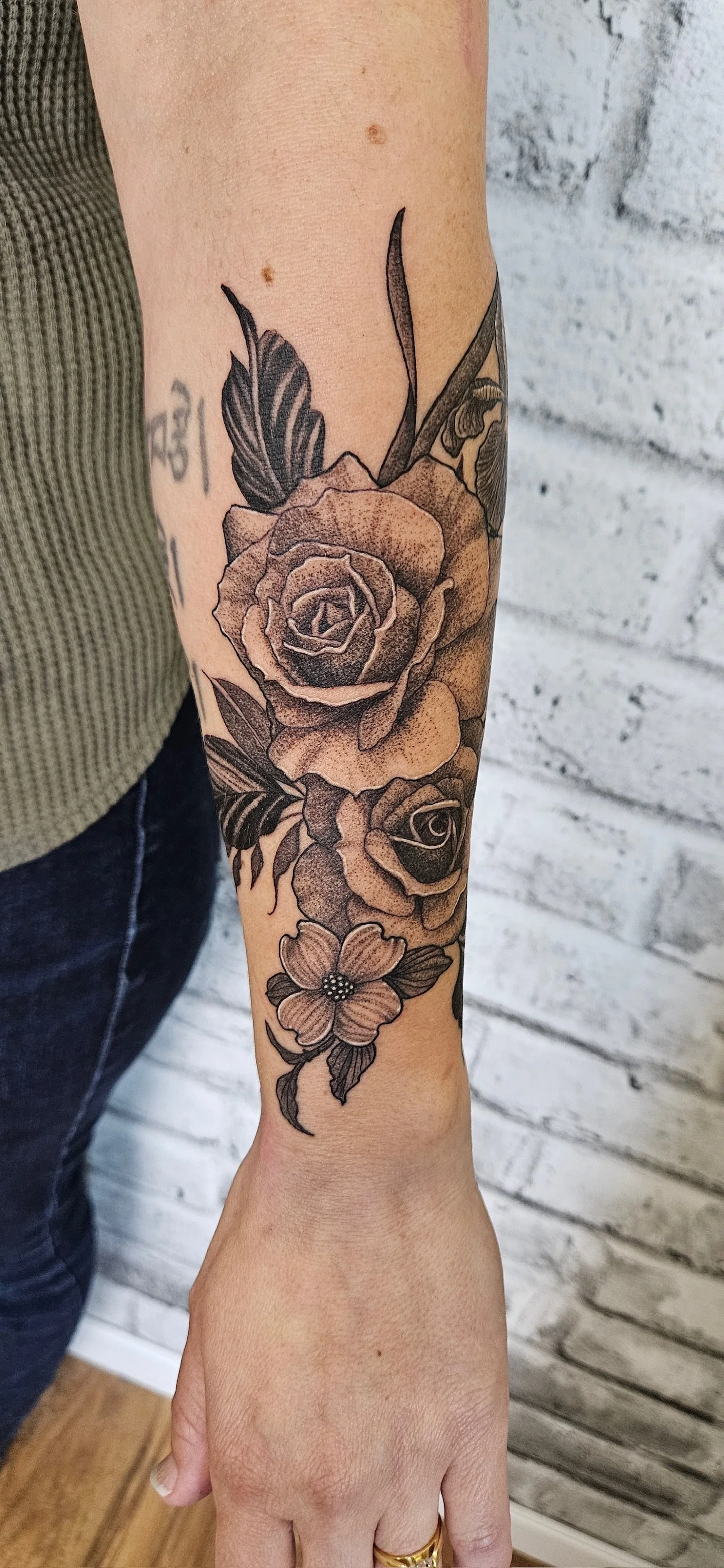 Black and gray floral tattoo featuring roses, a flower, and leaves on a person's forearm. Fine line delicate feminine flowers.