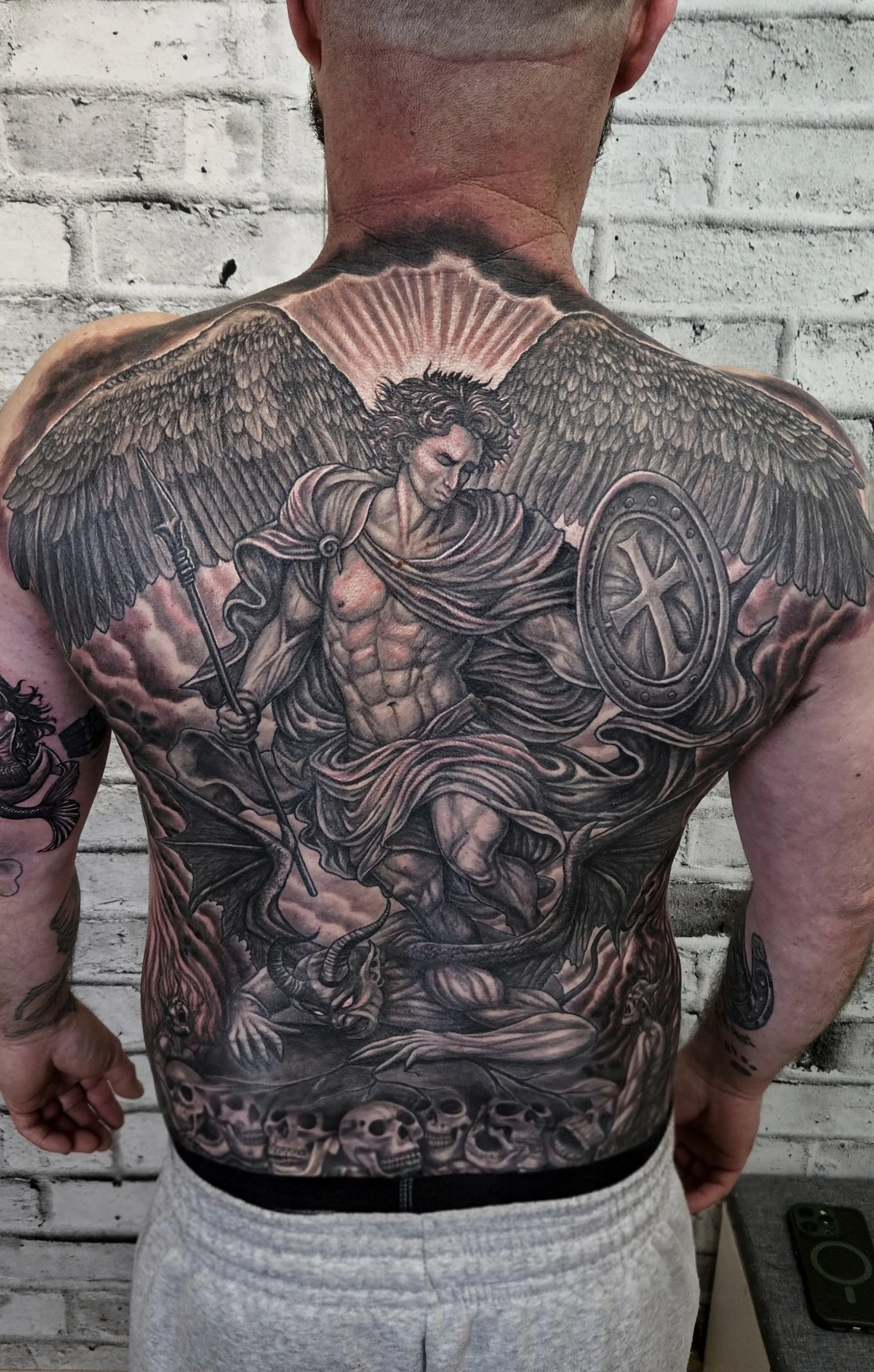 A detailed black and gray Backpeice tattoo of a muscular angel warrior with large wings, a shield with a cross, and a spear, covering the chest and abdomen, against a background of a skull and clouds. Arc Angel Gabriel crushing the devil. 